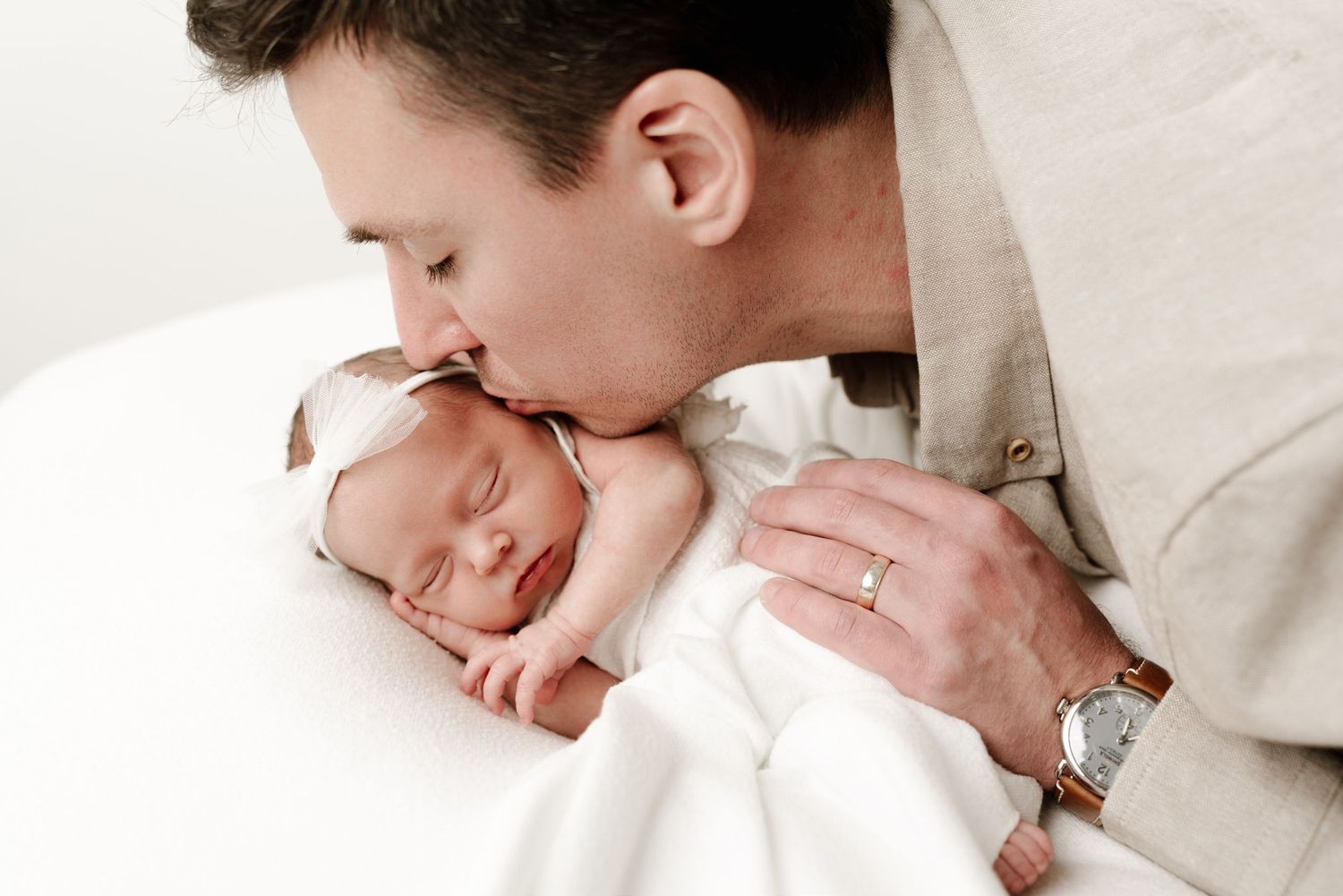 A father gently kisses his sleeping newborn baby wrapped in white, capturing a tender bonding moment.