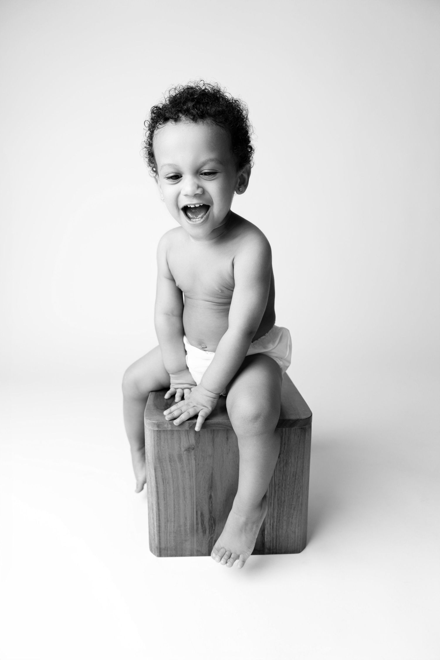 Black and white photo of a laughing baby in a diaper sitting on a wooden block against a white background.