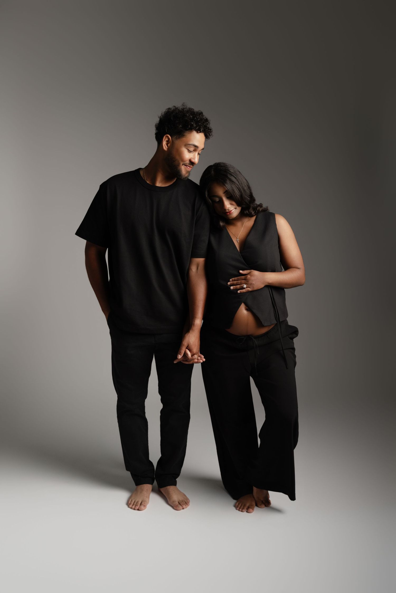 A pregnant couple dressed in black holding hands, the woman cradling her baby bump in a studio setting.