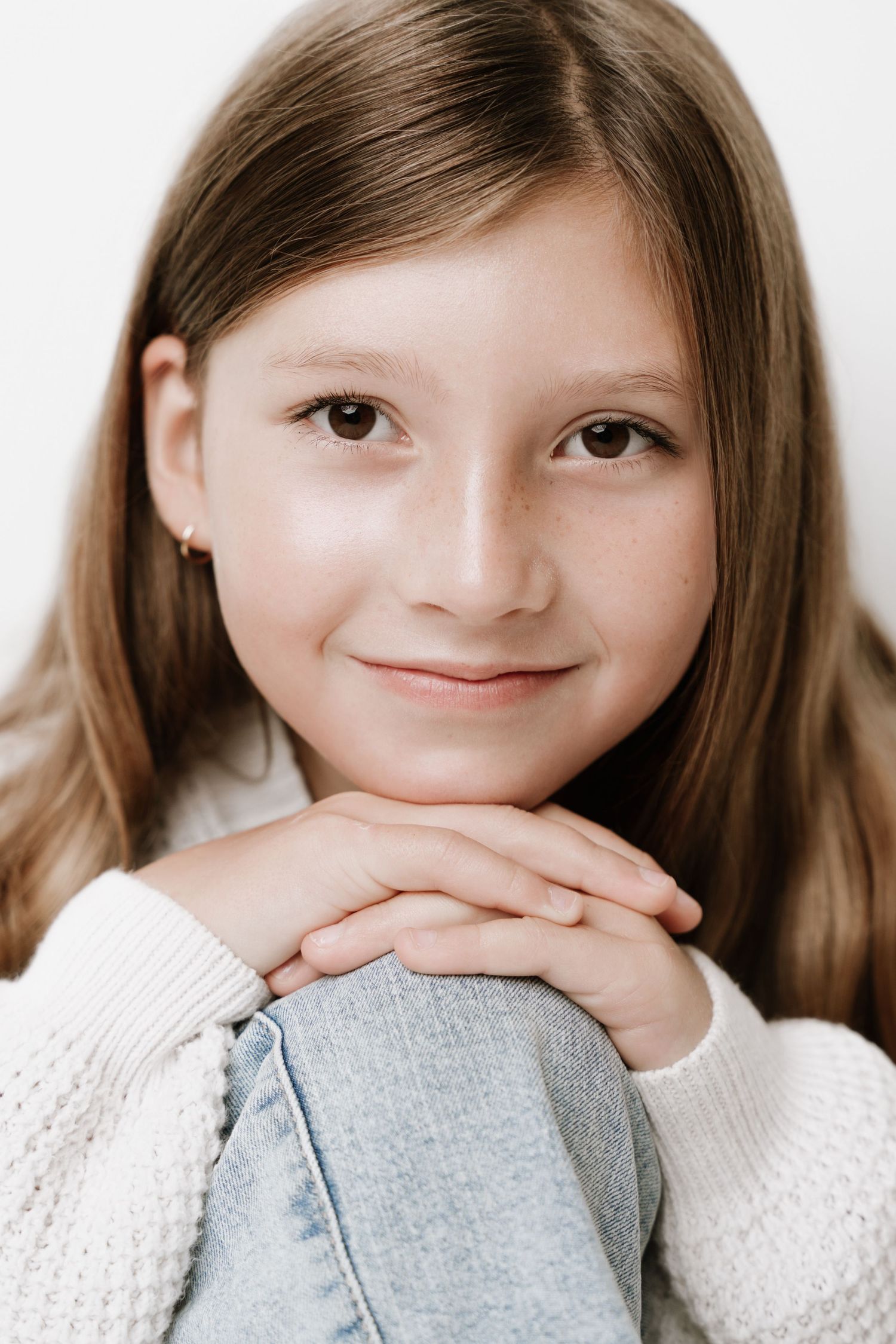 A young girl with brown hair smiles warmly, resting her chin on clasped hands while wearing a white sweater.