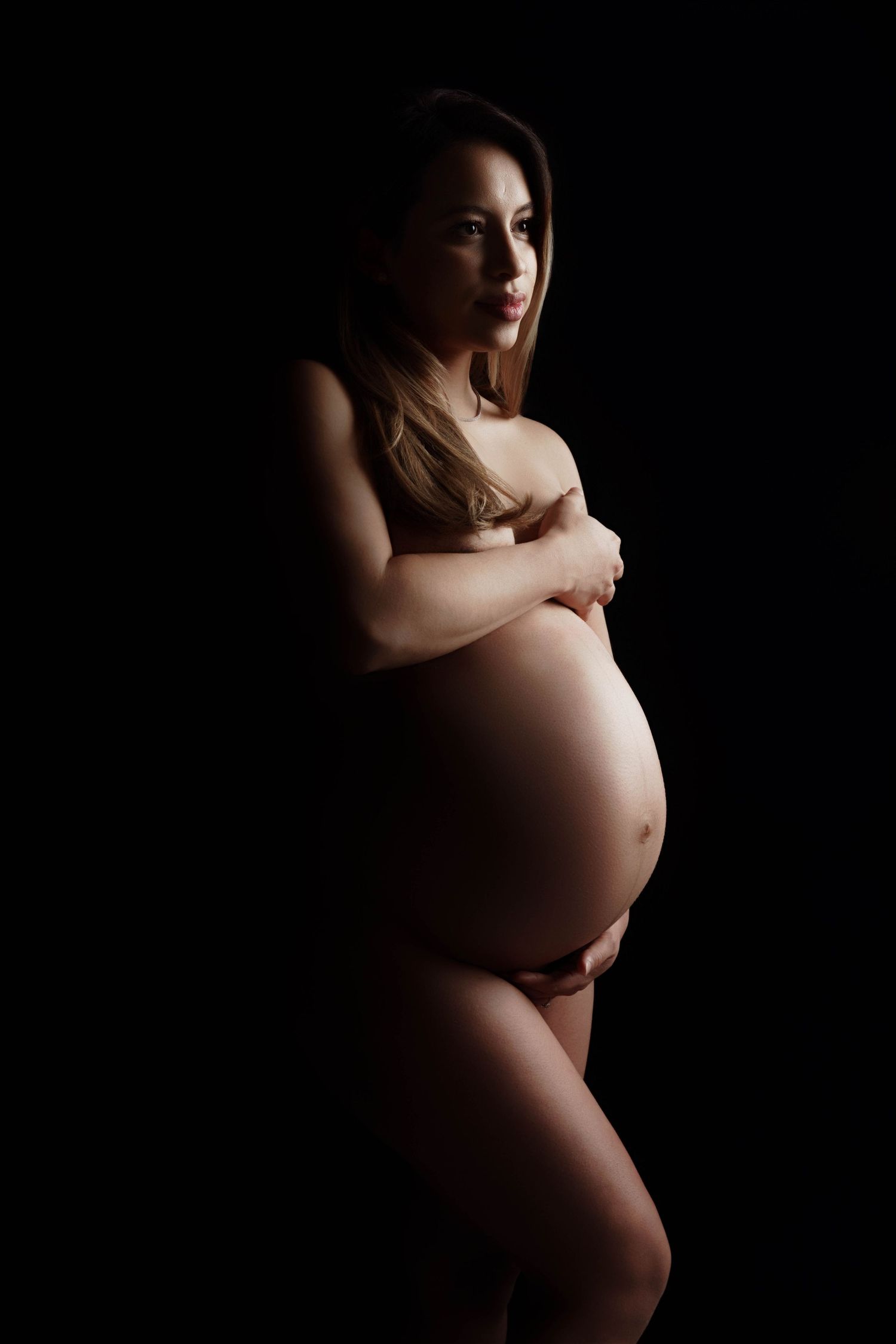 Dramatic low-key maternity portraits highlighting the pregnant silhouette against dark background.