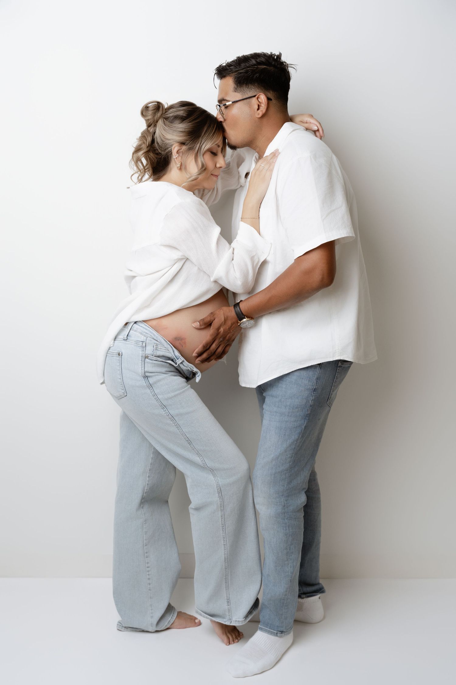 A couple in matching white shirts and light jeans embraces against a white wall in a series of romantic poses.