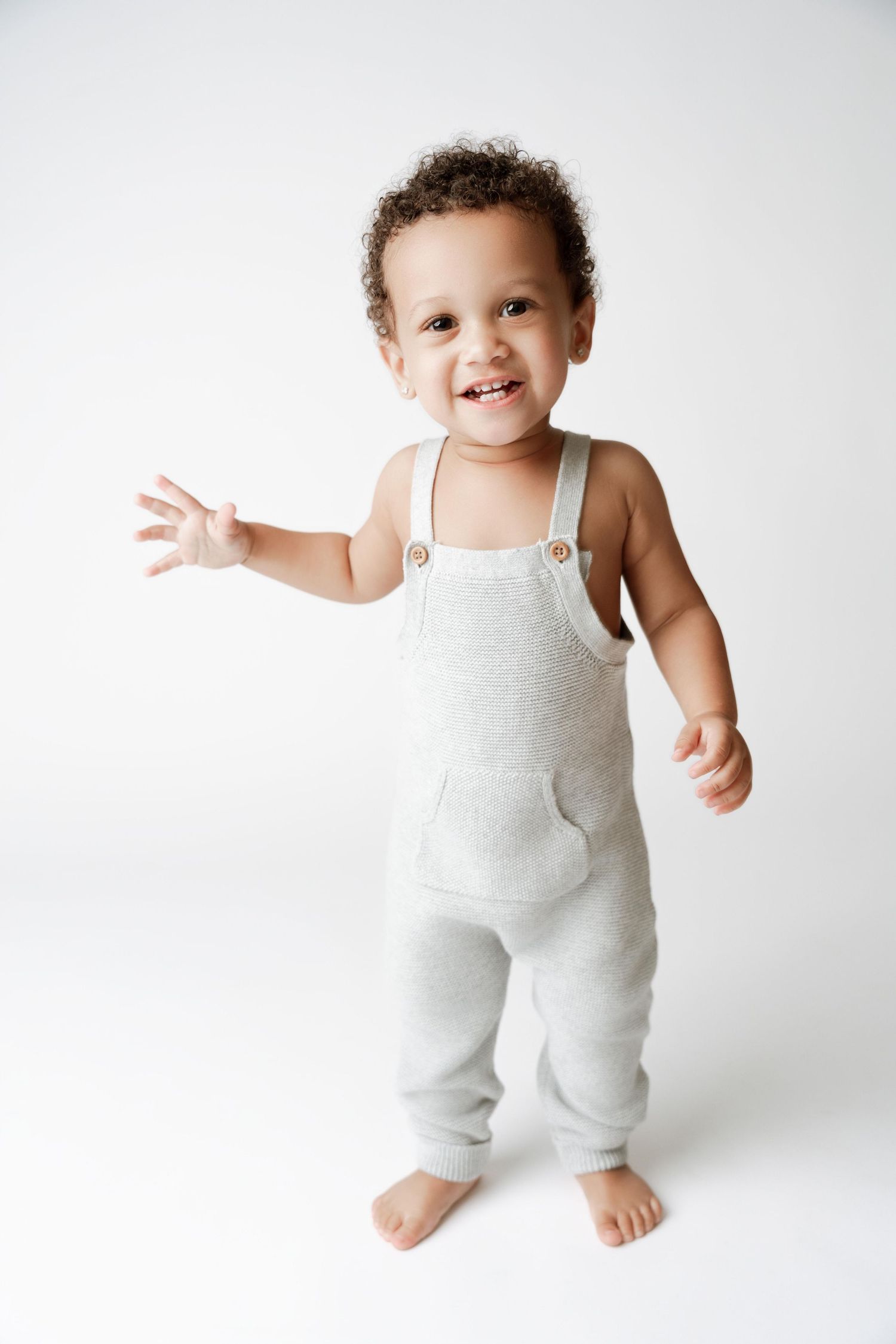 Smiling toddler in gray overalls standing barefoot on white background, waving hand cheerfully.