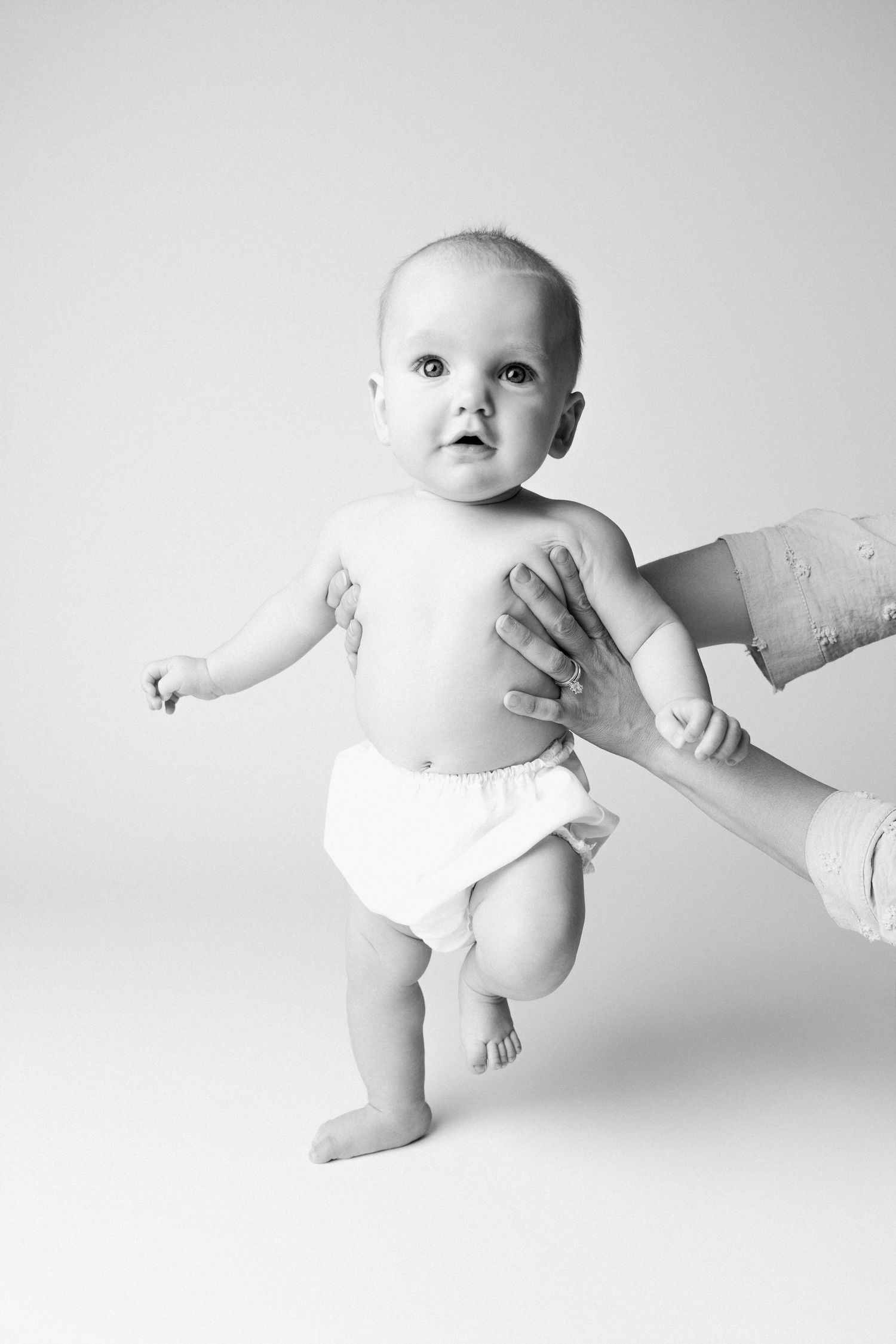 Black and white photo of a baby in a diaper standing upright with adult hands providing gentle support.