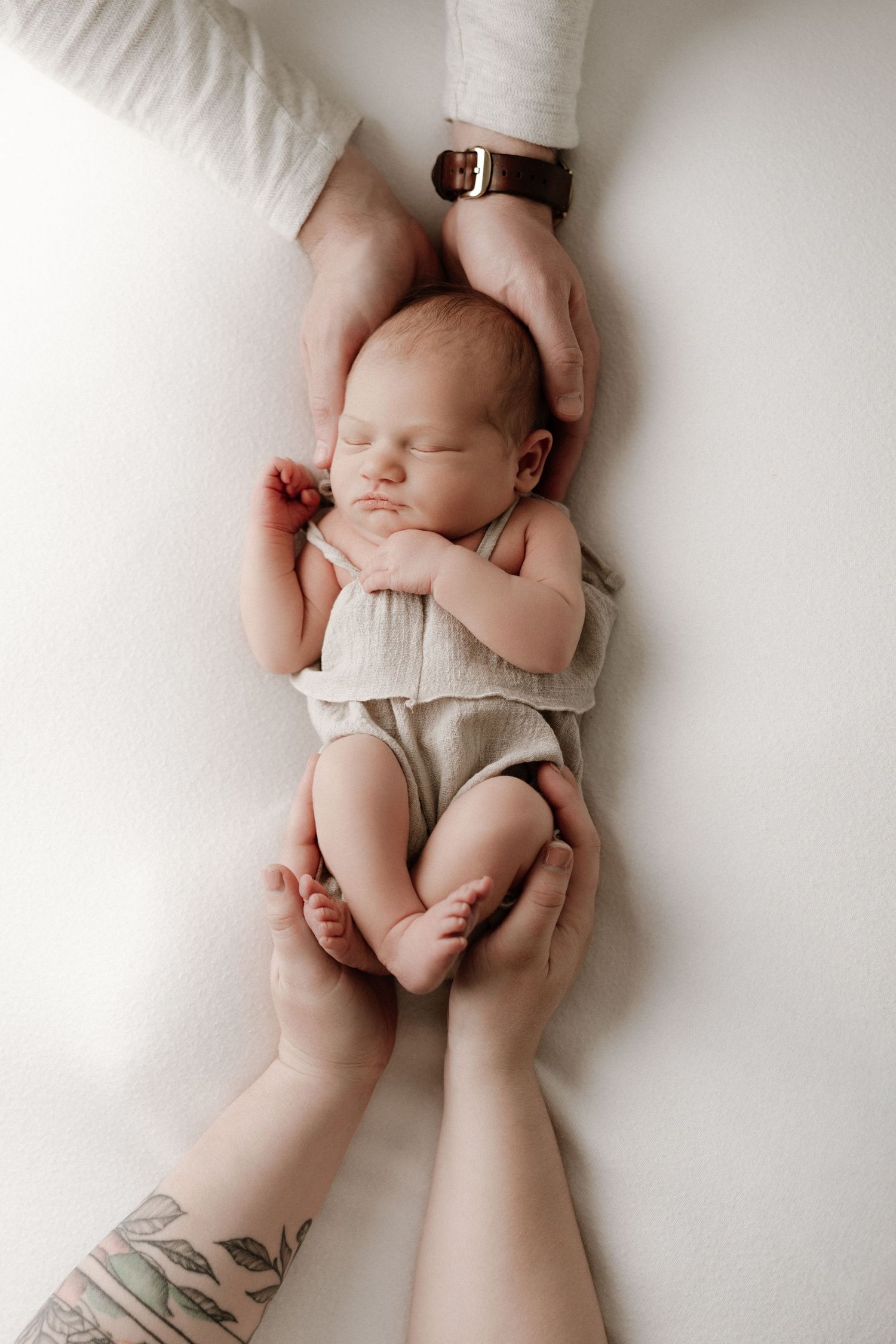 Sleeping newborn baby cradled by parents hands, wearing a neutral linen romper on a white background.