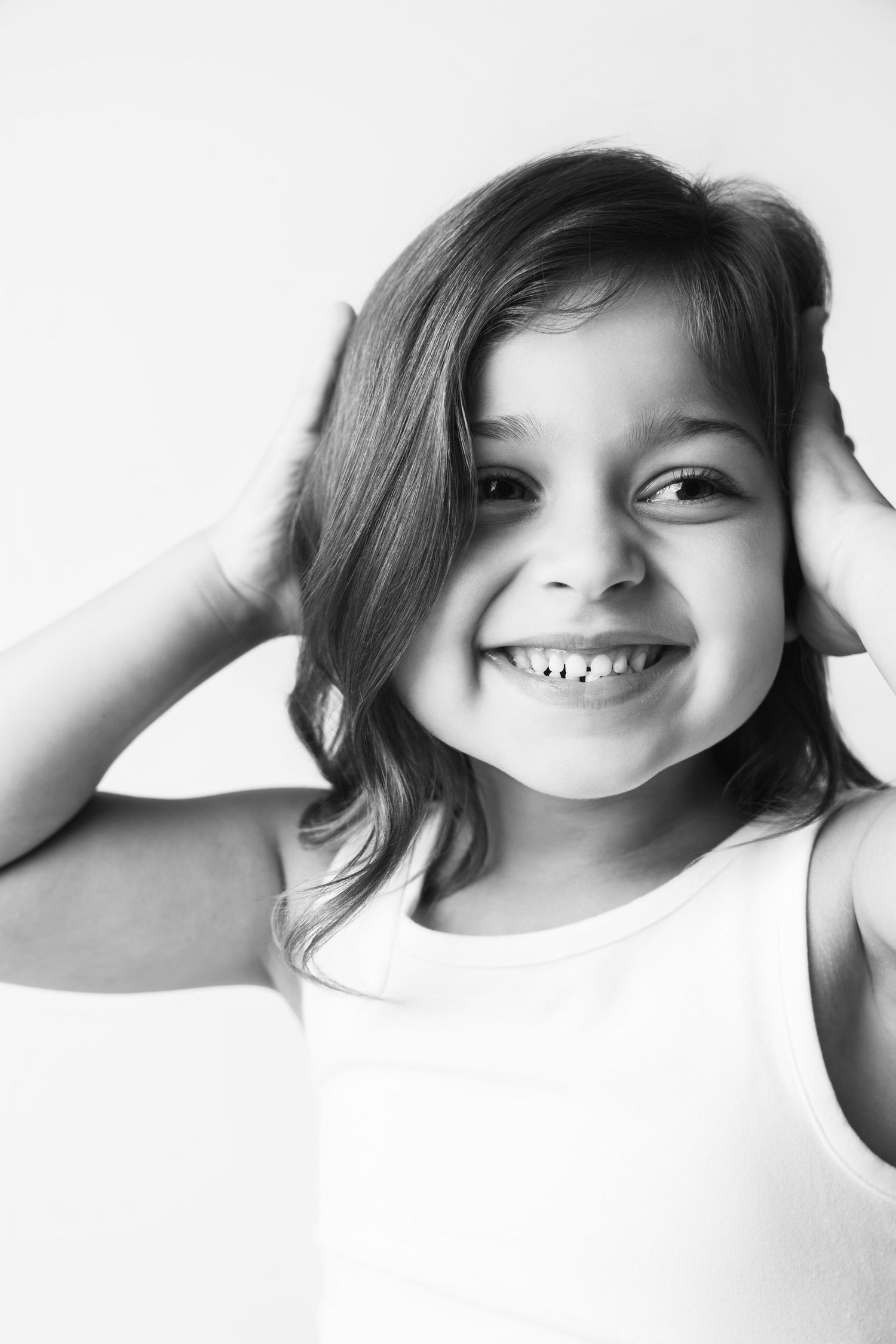 Black and white portrait of a smiling young girl with hands on head wearing a white tank top.