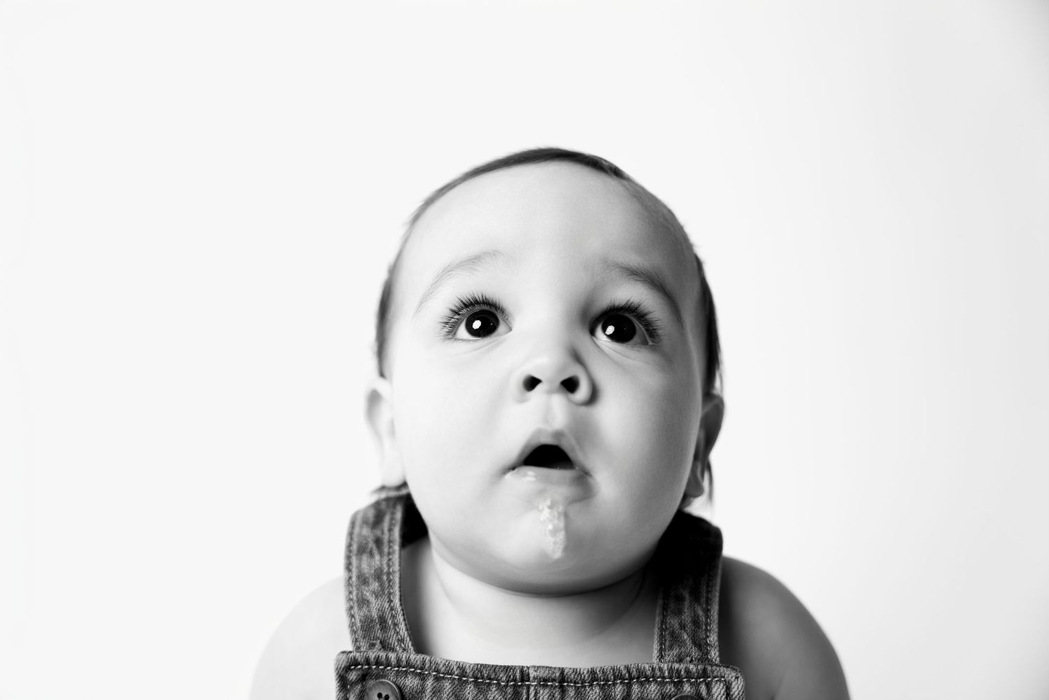 Black and white portrait of a cute baby with drool, looking up curiously, wearing denim overalls.