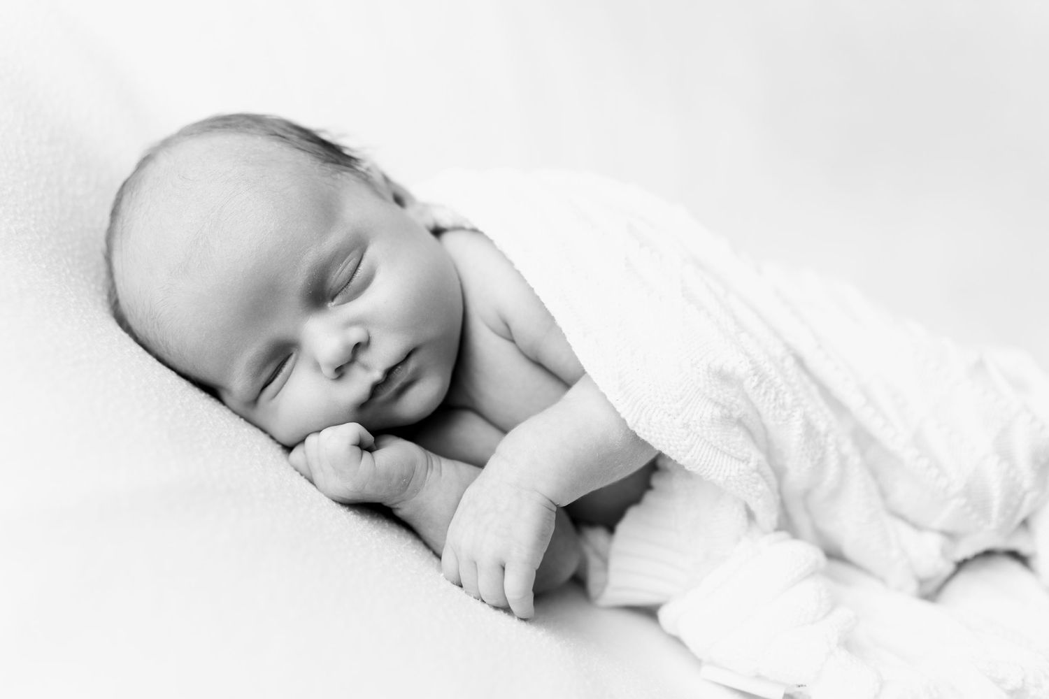 Black and white newborn baby sleeping peacefully, wrapped in soft white blanket, hands tucked under chin.