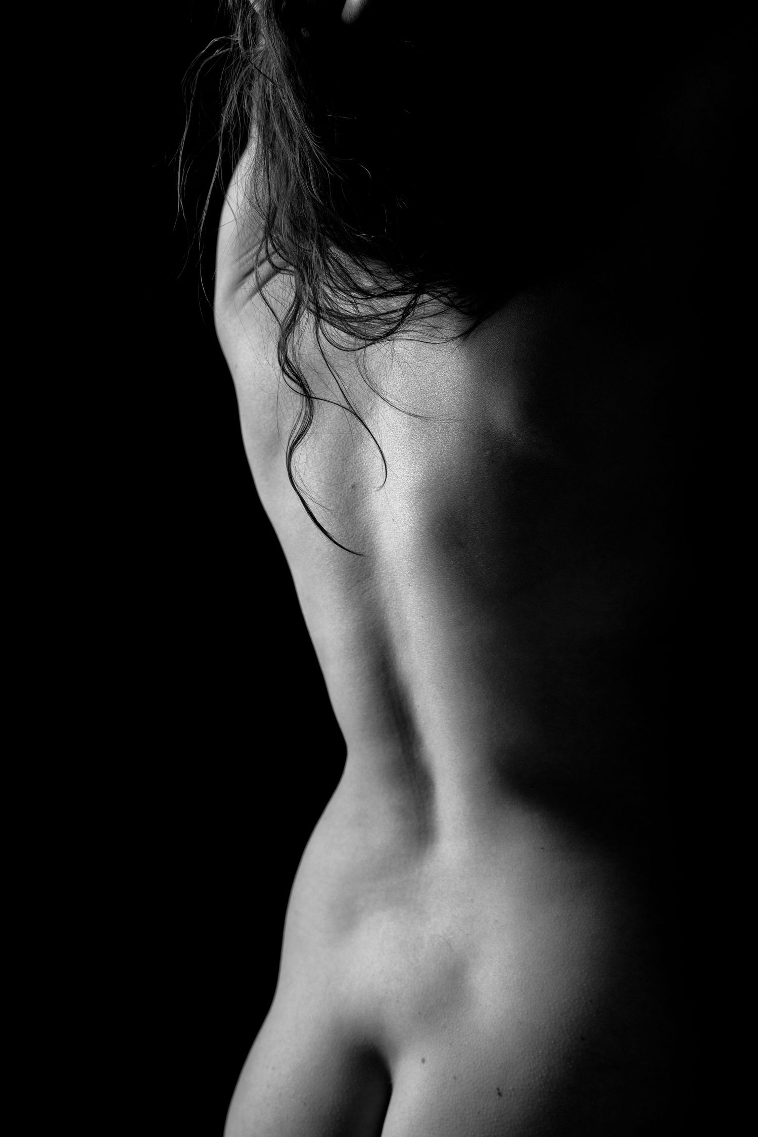 Black and white artistic nude photography highlighting the graceful curves of a woman's back against dark background.
