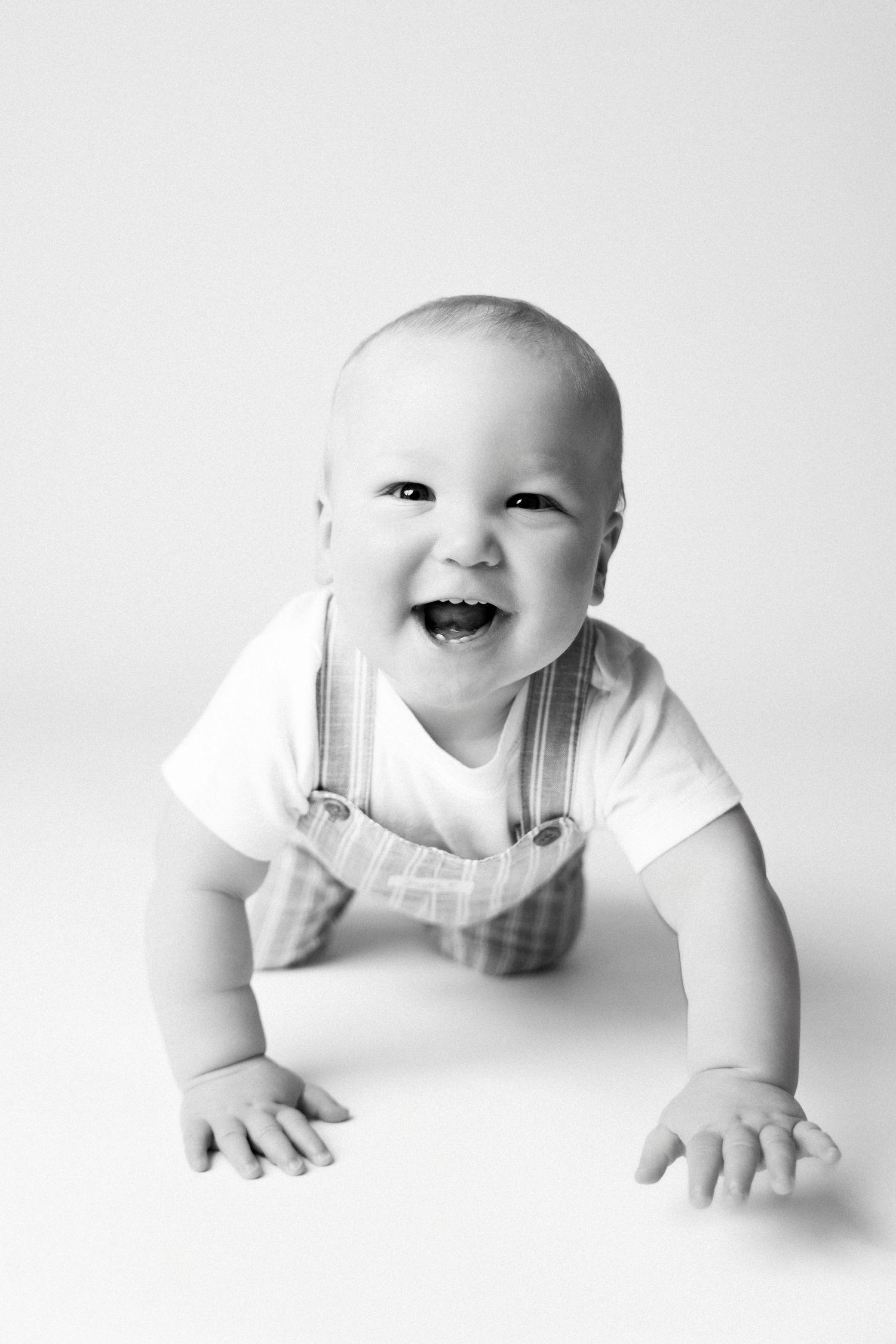 Black and white photo of a happy smiling baby in overalls crawling on a white background.