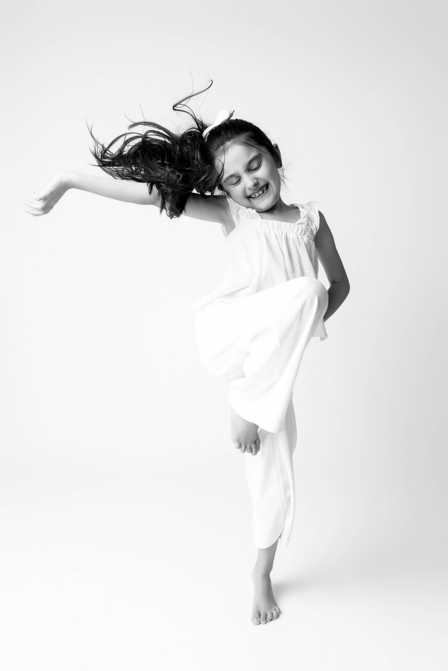 A joyful young girl in a white dress dances barefoot, hair flying, in a black and white studio photo.