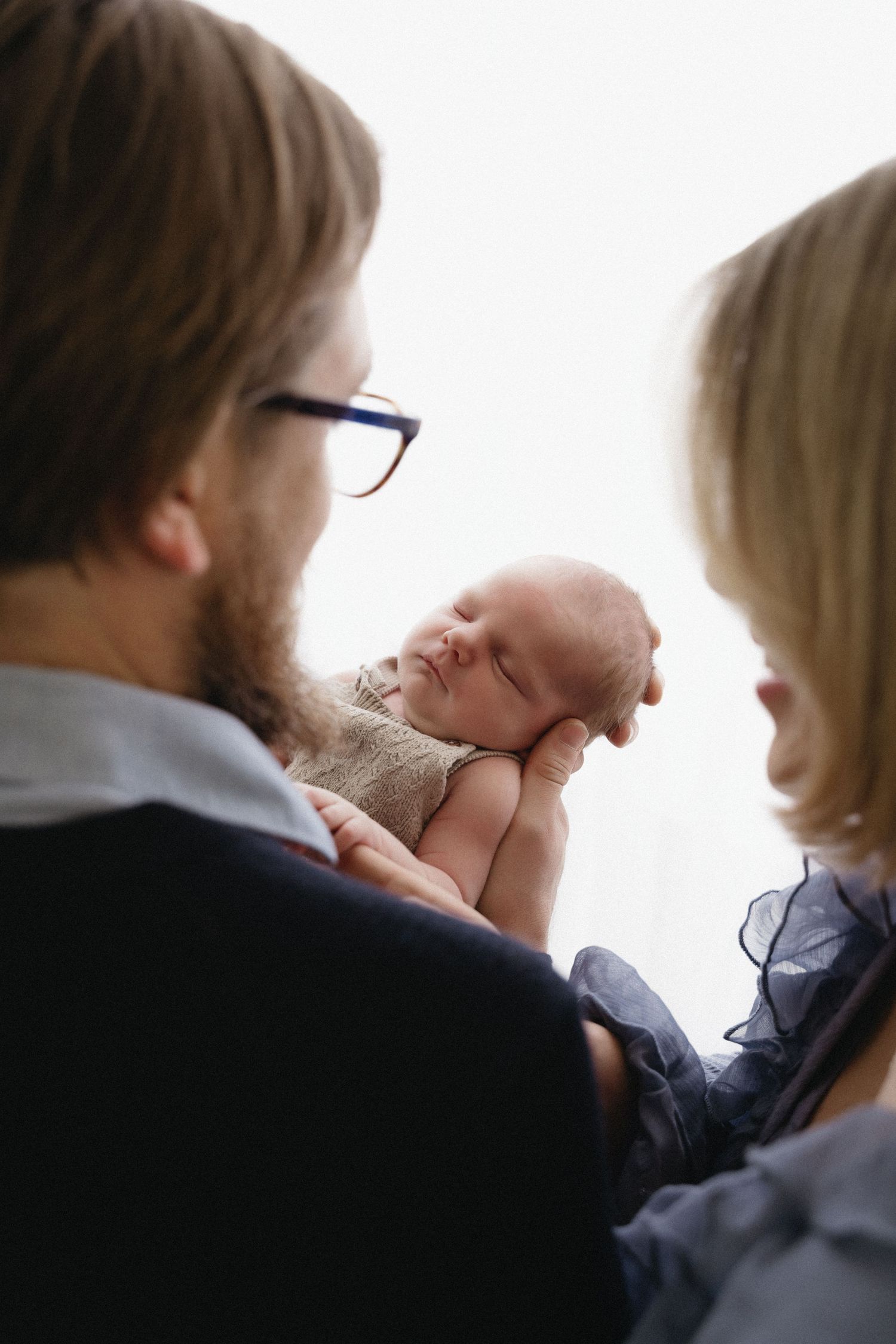 A tender family moment shows parents gazing lovingly at their newborn baby against a white background.
