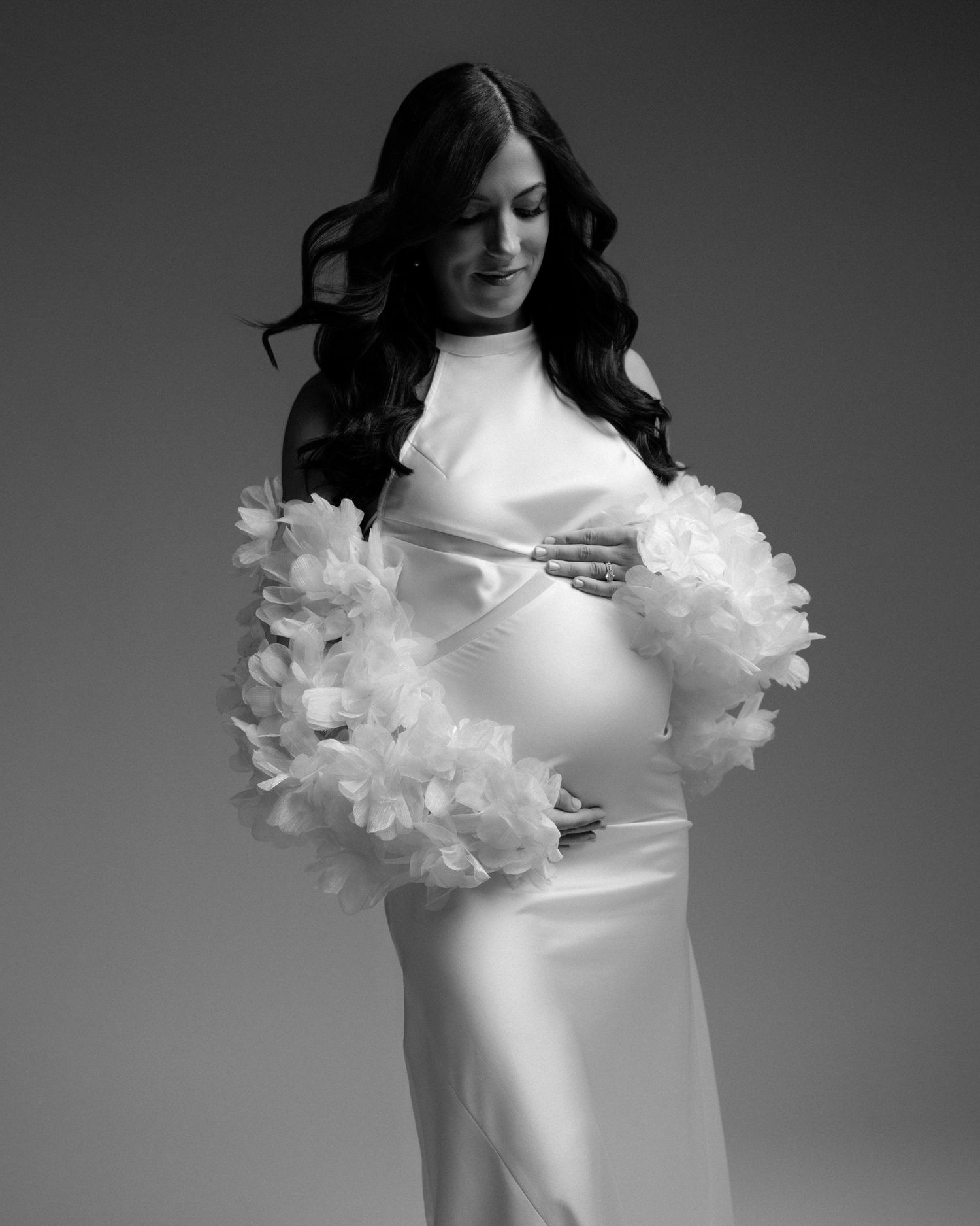 Pregnant woman in white satin gown with floral feather sleeves, cradling baby bump in elegant black and white maternity photo.