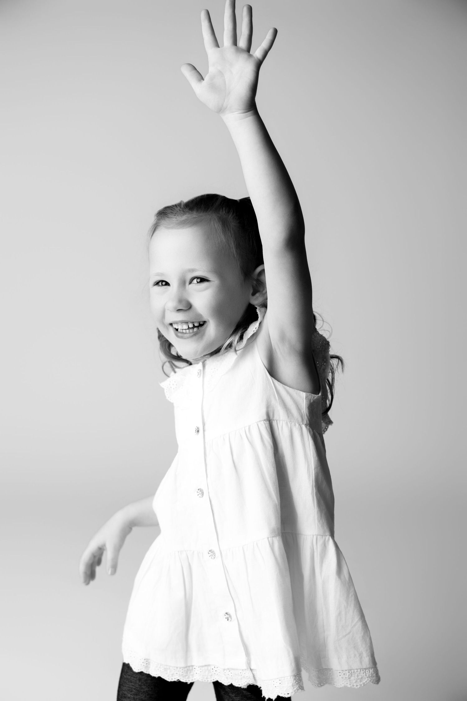 Energetic child in white top raising arms and jumping joyfully against grey background.