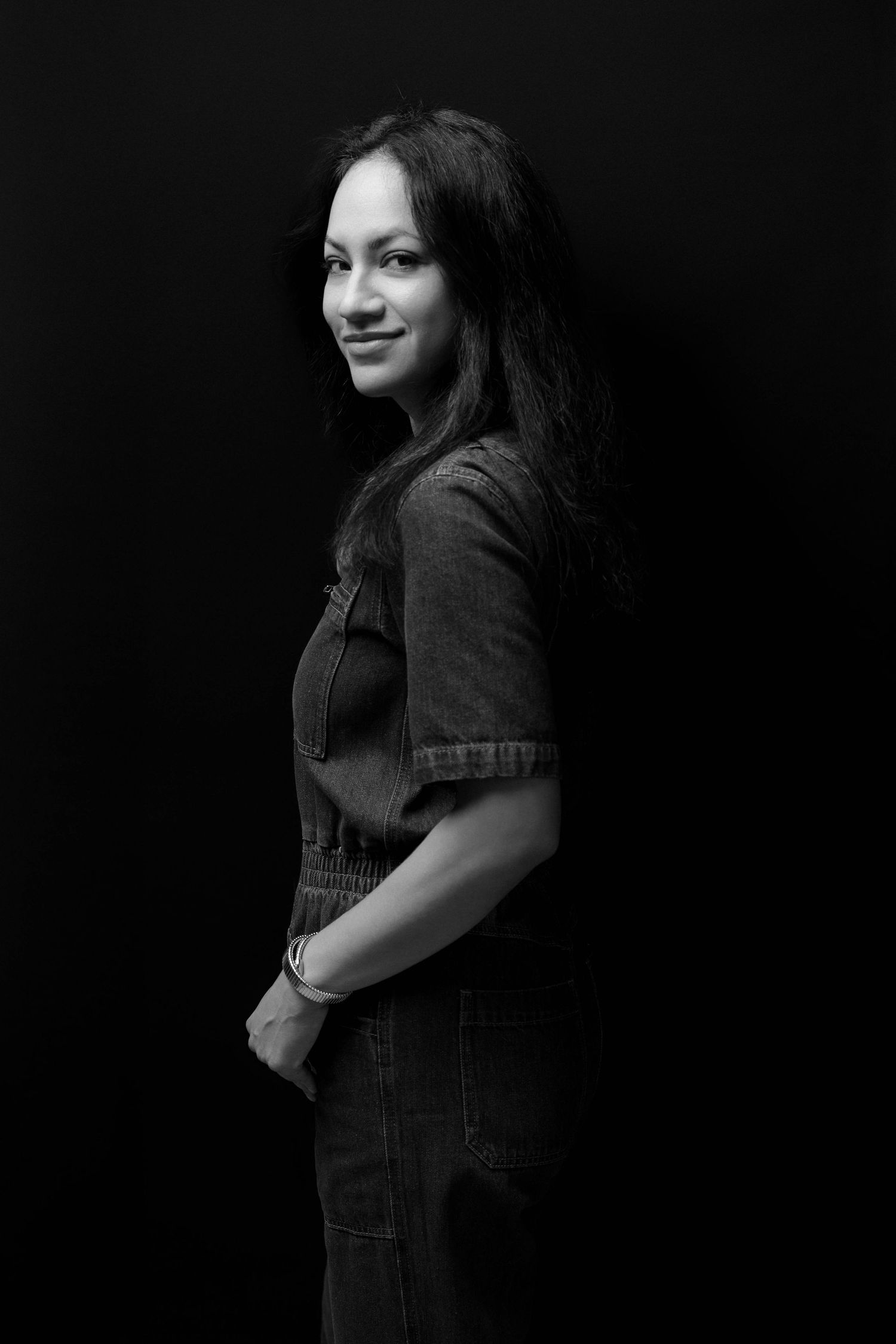 Black and white portrait of a young woman in denim overalls smiling over her shoulder against a dark background.