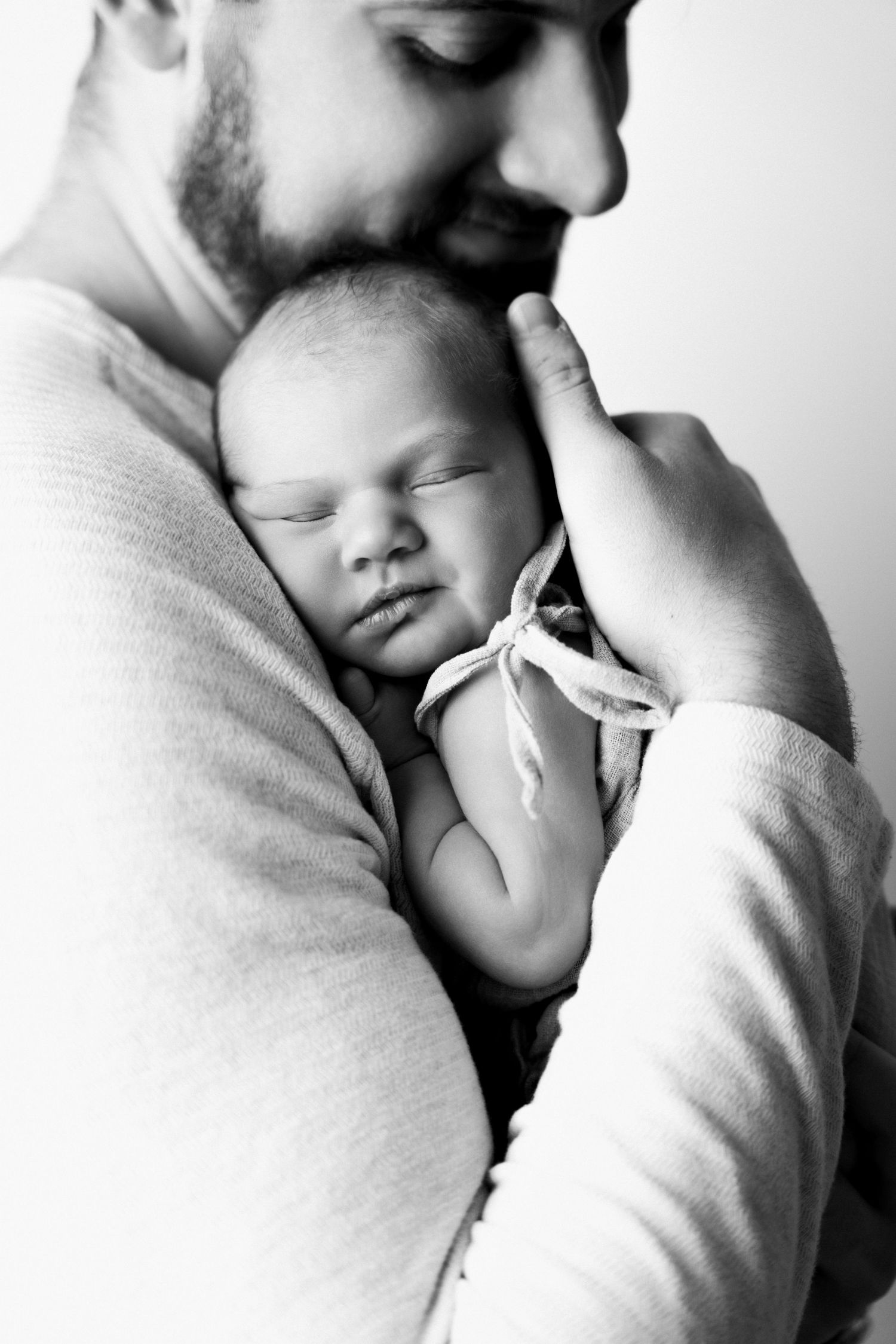 Black and white photo of a bearded father tenderly holding his sleeping newborn baby close to his chest.