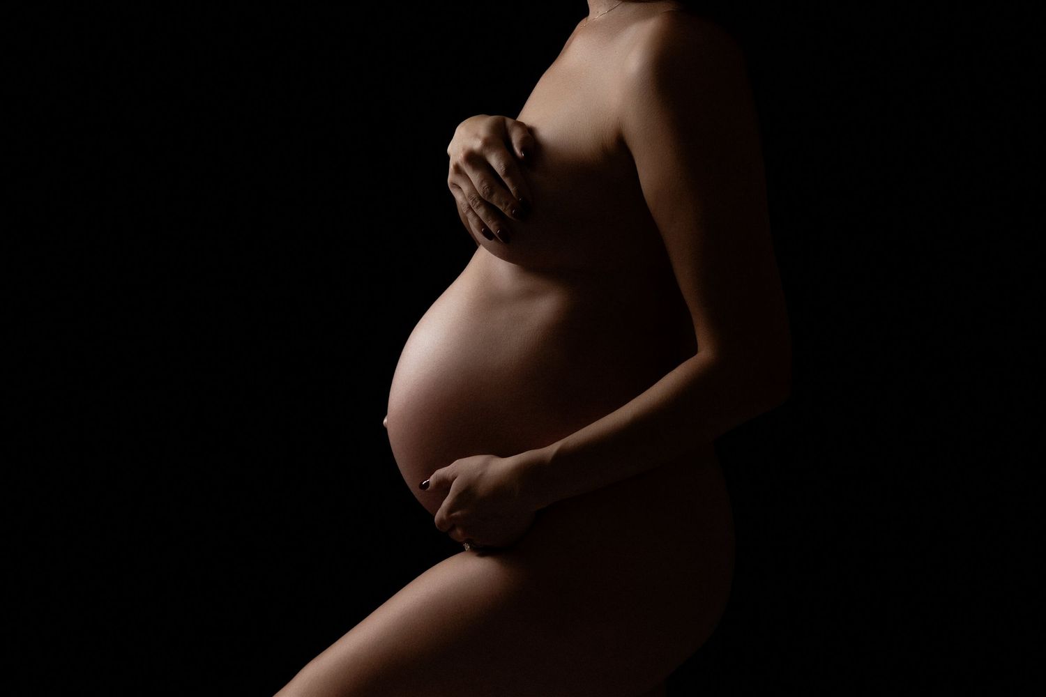 Artistic silhouette maternity photo with dramatic lighting showing pregnant belly against dark background.