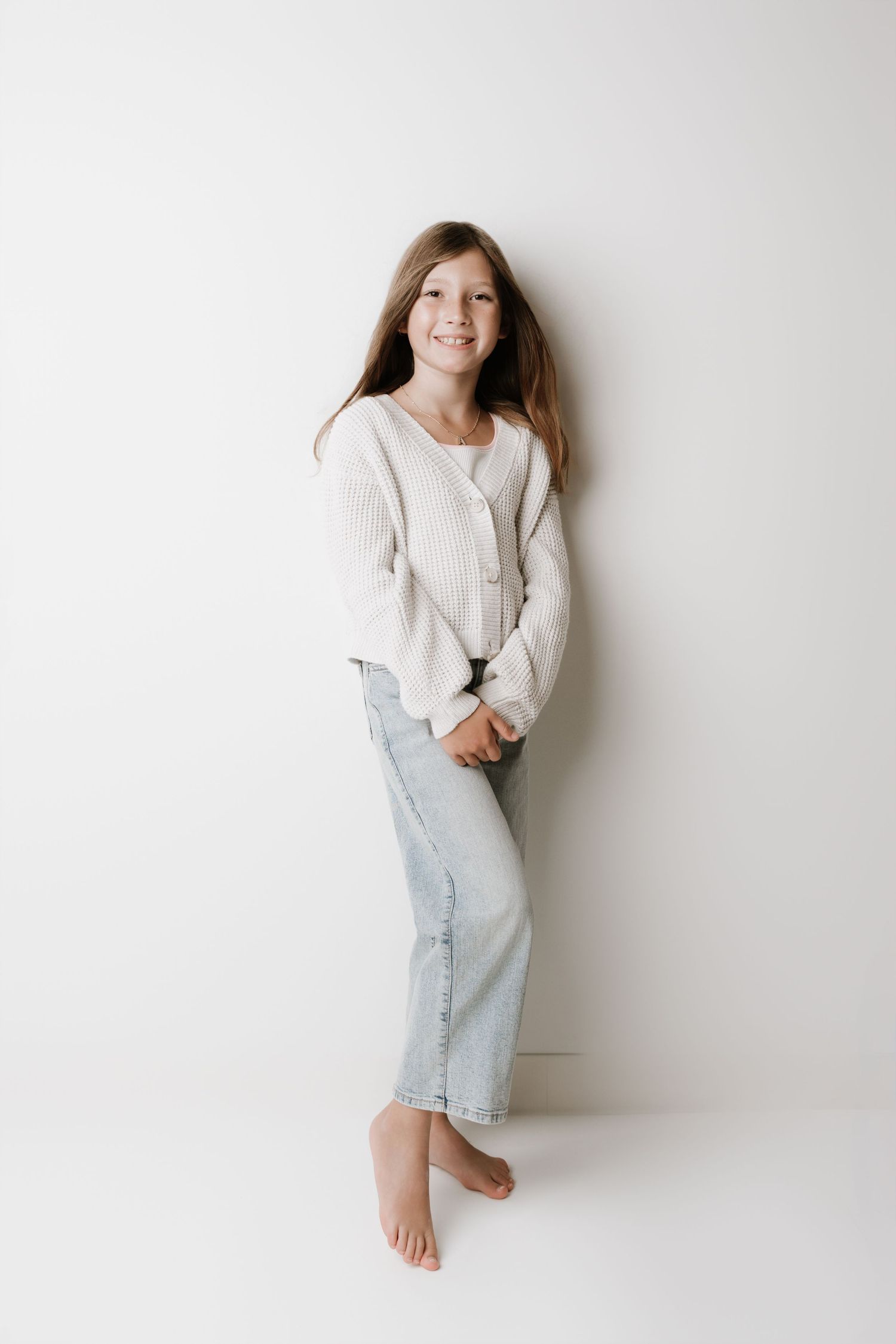 Smiling young girl in white cardigan and light jeans standing barefoot against a white wall.