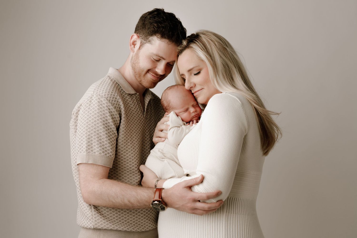 A couple tenderly cradles their newborn baby in a soft, neutral-toned studio portrait.