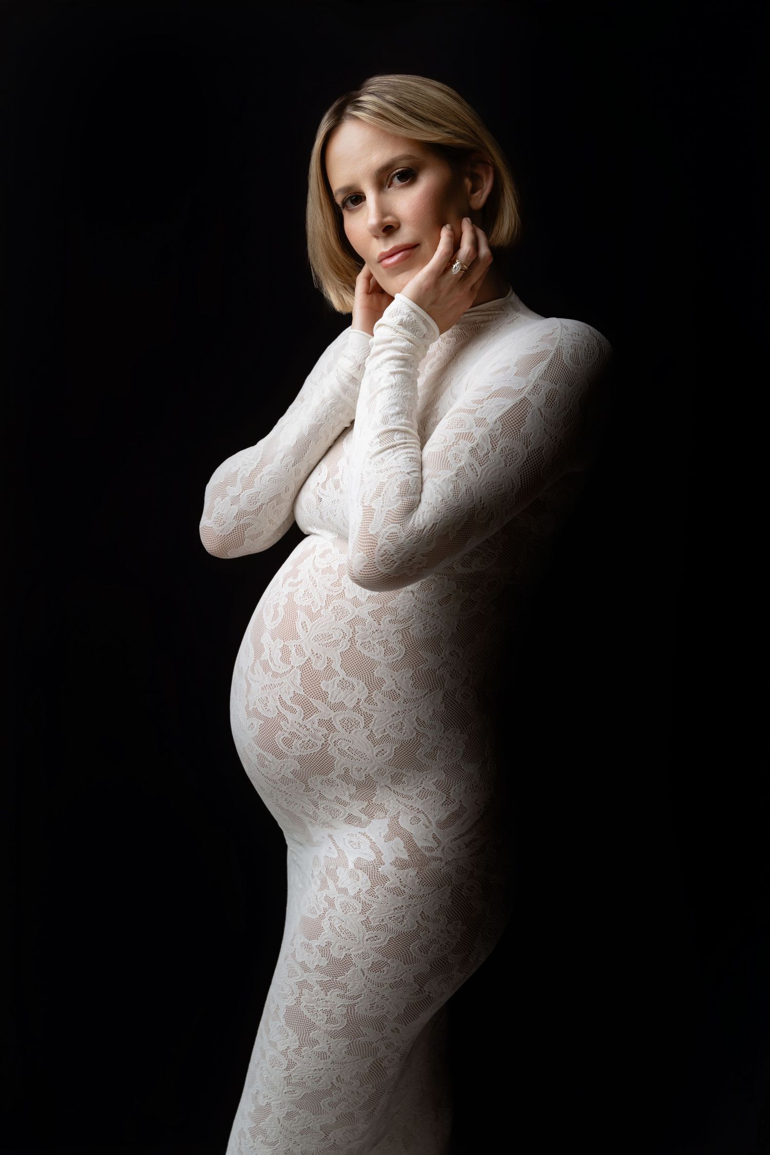 Elegant maternity portrait featuring a white lace dress against a dark background in dramatic lighting.