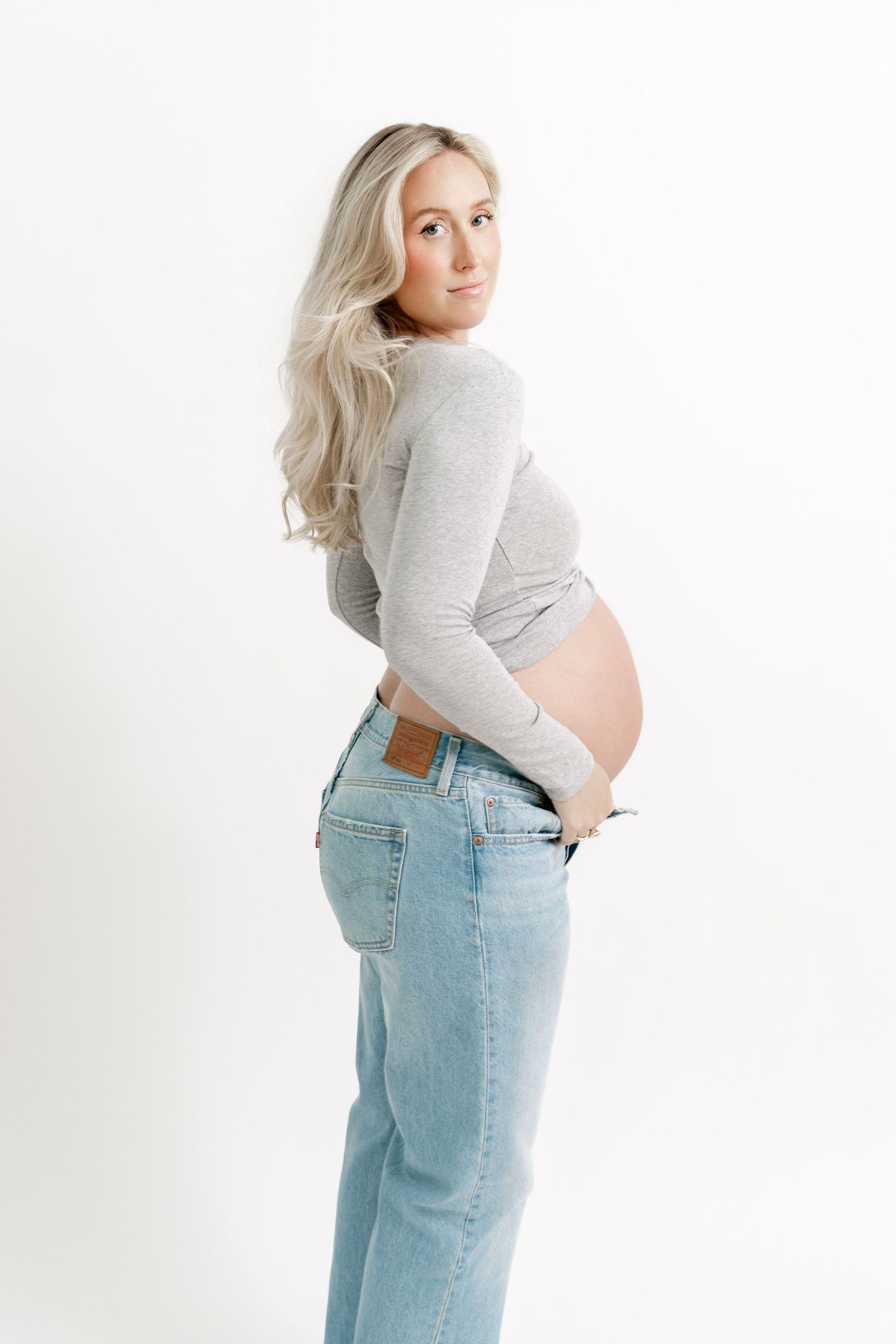 A pregnant woman in a grey sweater and light wash jeans poses sideways against a white backdrop.