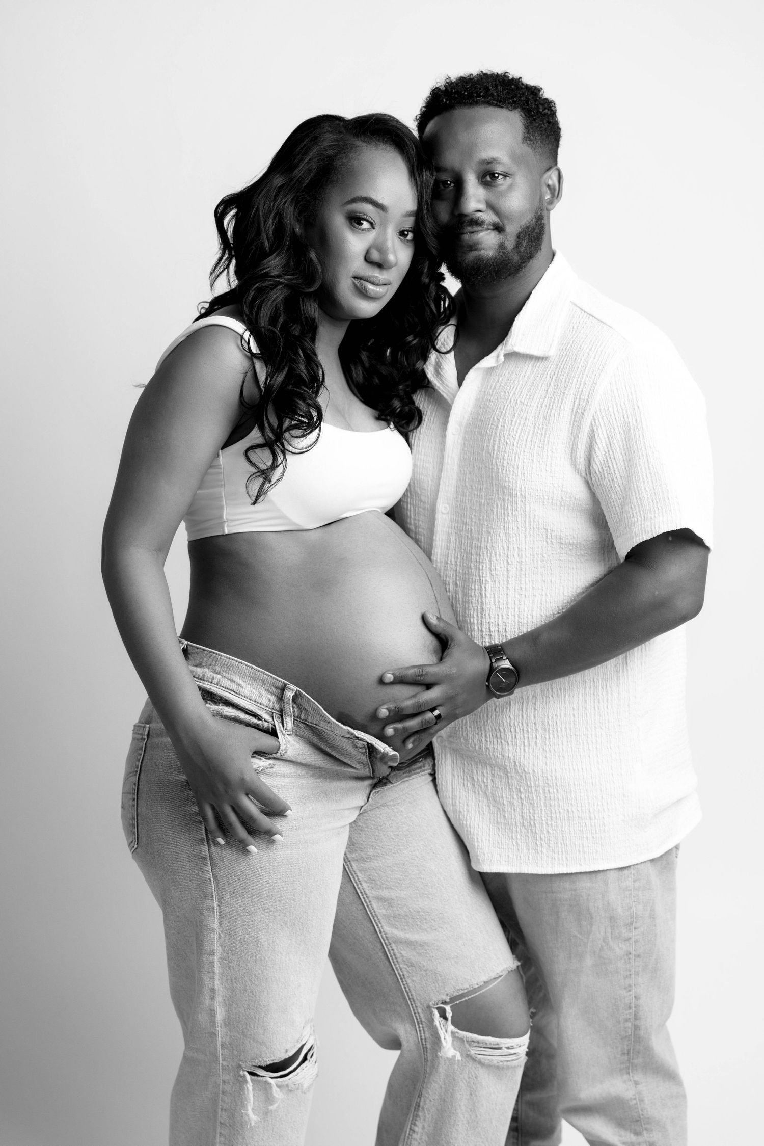Intimate black and white maternity photo series showing a couple in white outfits embracing against studio backdrop.