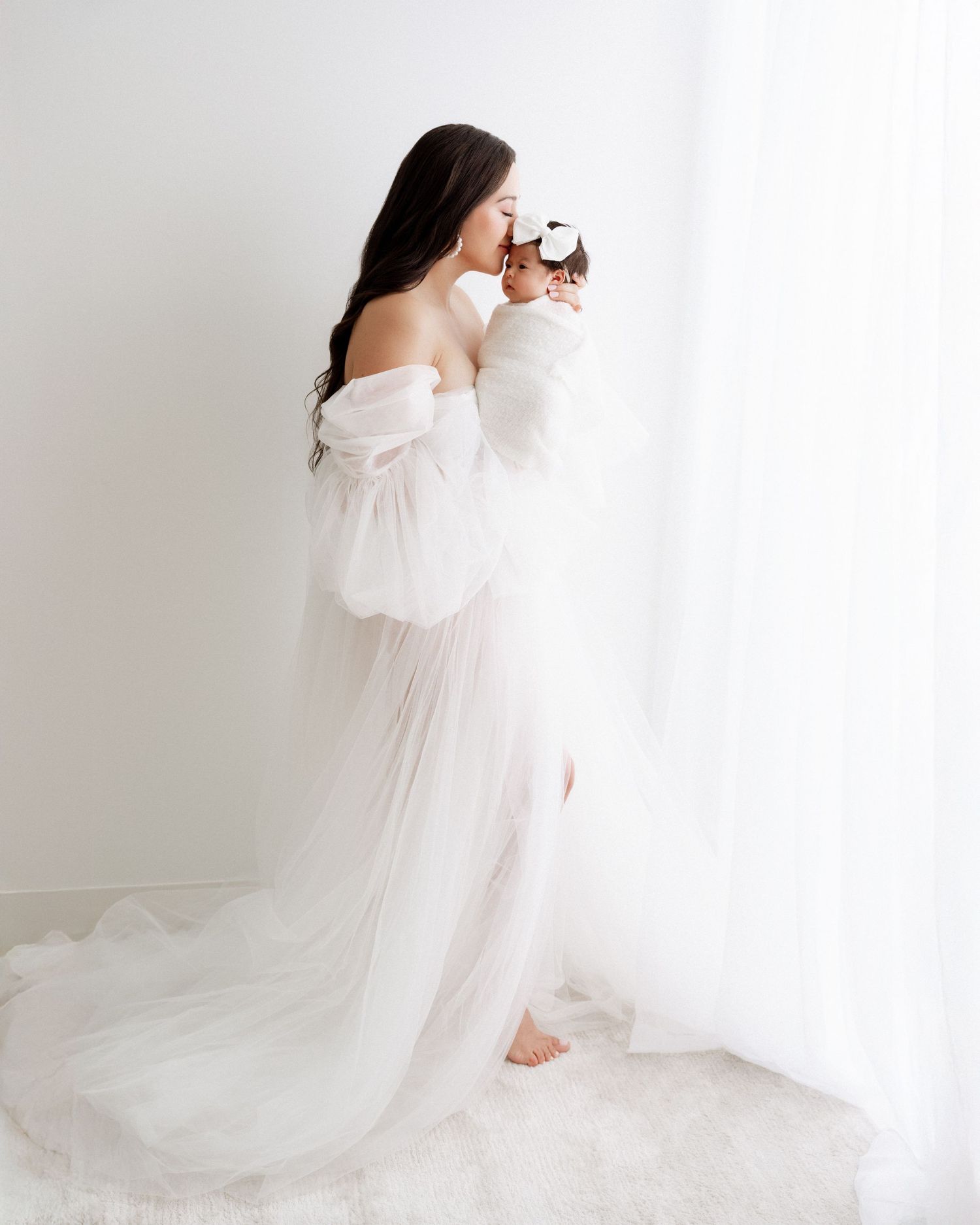 A woman in a flowing white maternity gown cradles her belly in a bright minimalist studio setting.