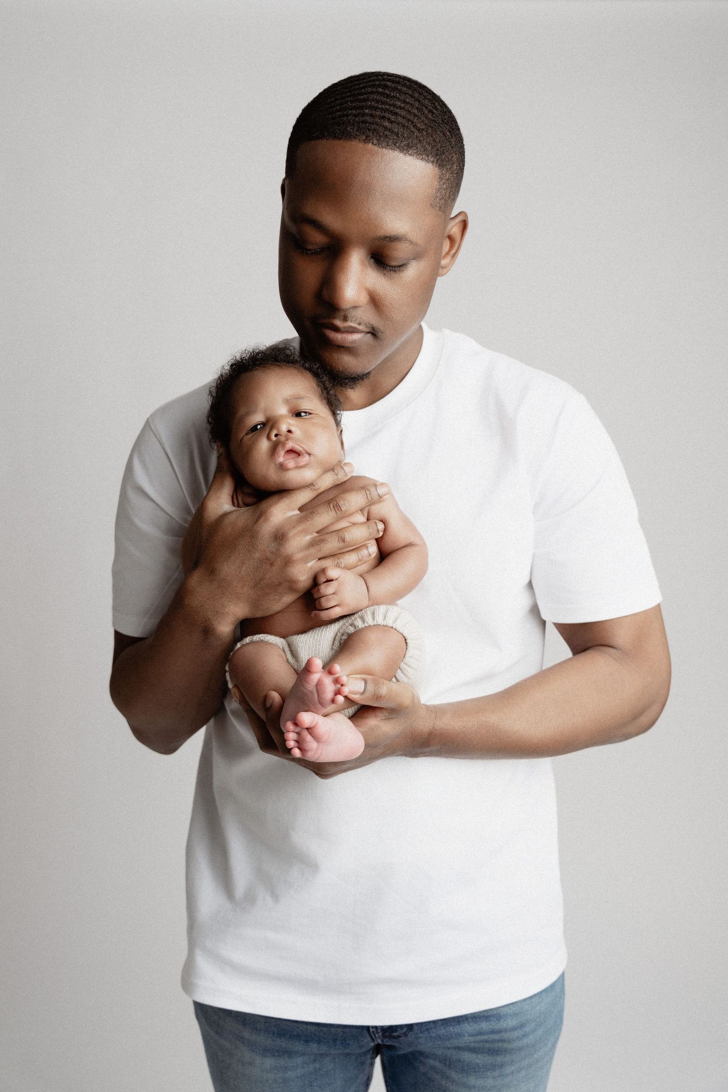 A parent in a white t-shirt tenderly holds their newborn baby against a light gray background.