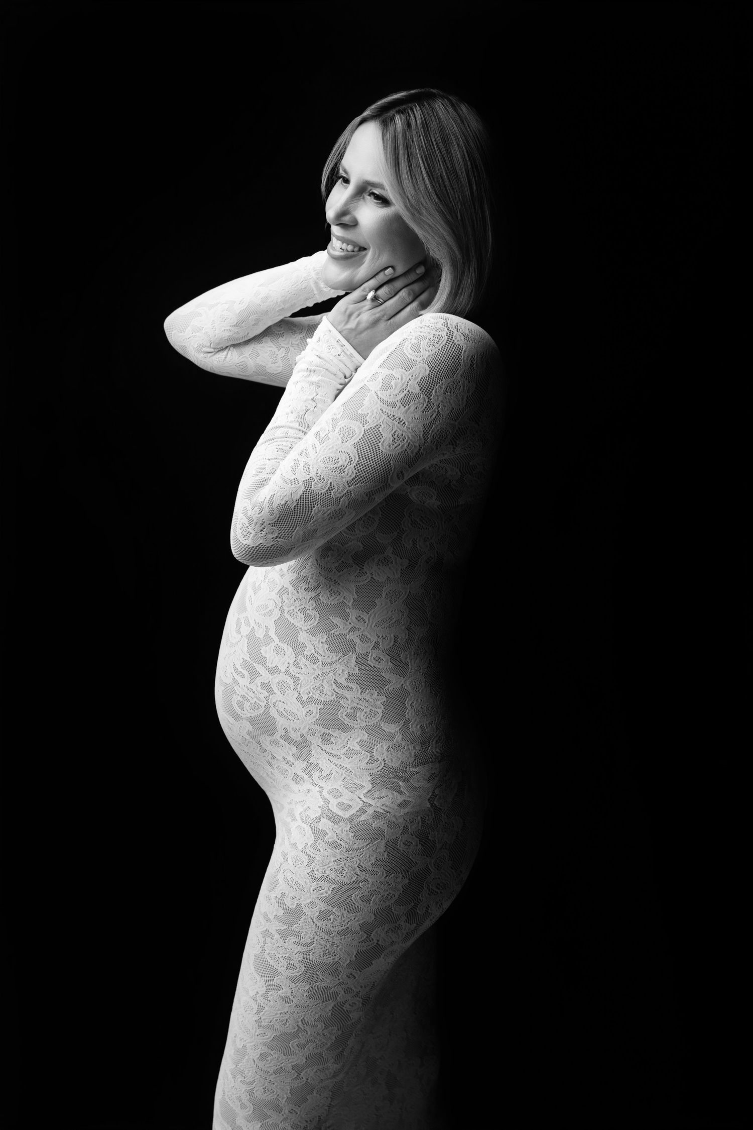 Black and white artistic maternity portrait of a woman in a lace dress against a dark background.