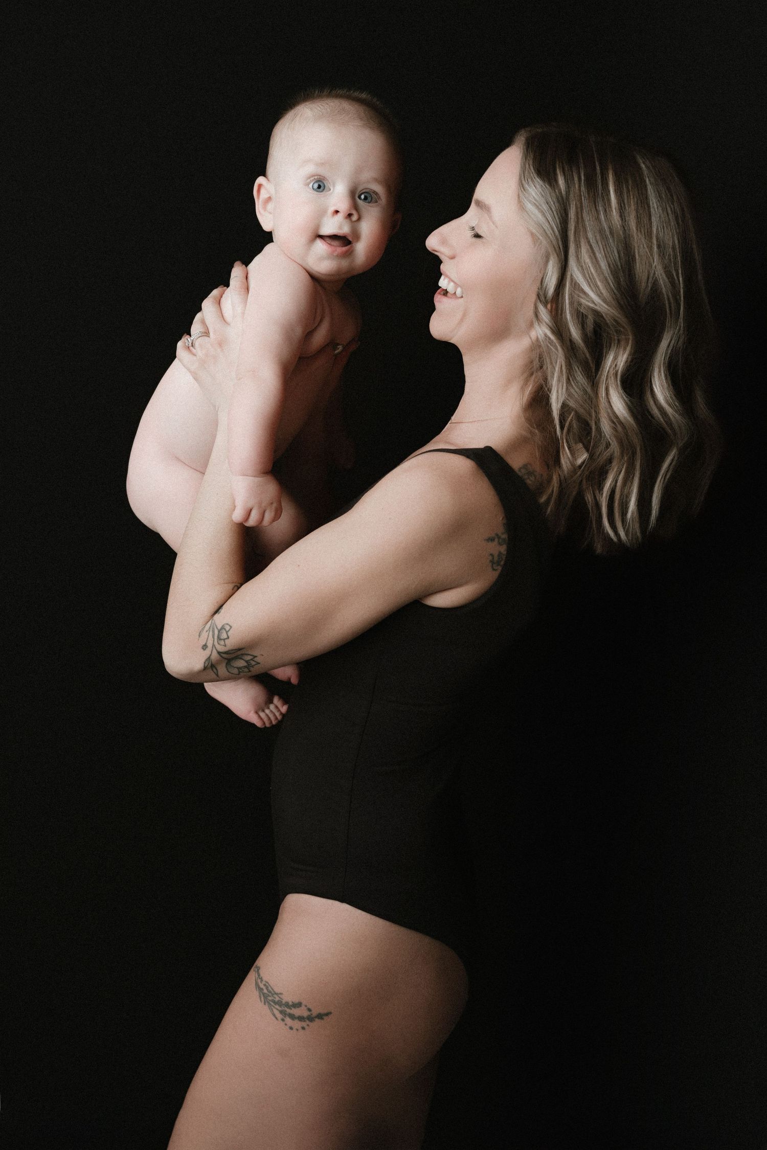A tender moment captured against a black backdrop as a woman in a black bodysuit holds a smiling baby.