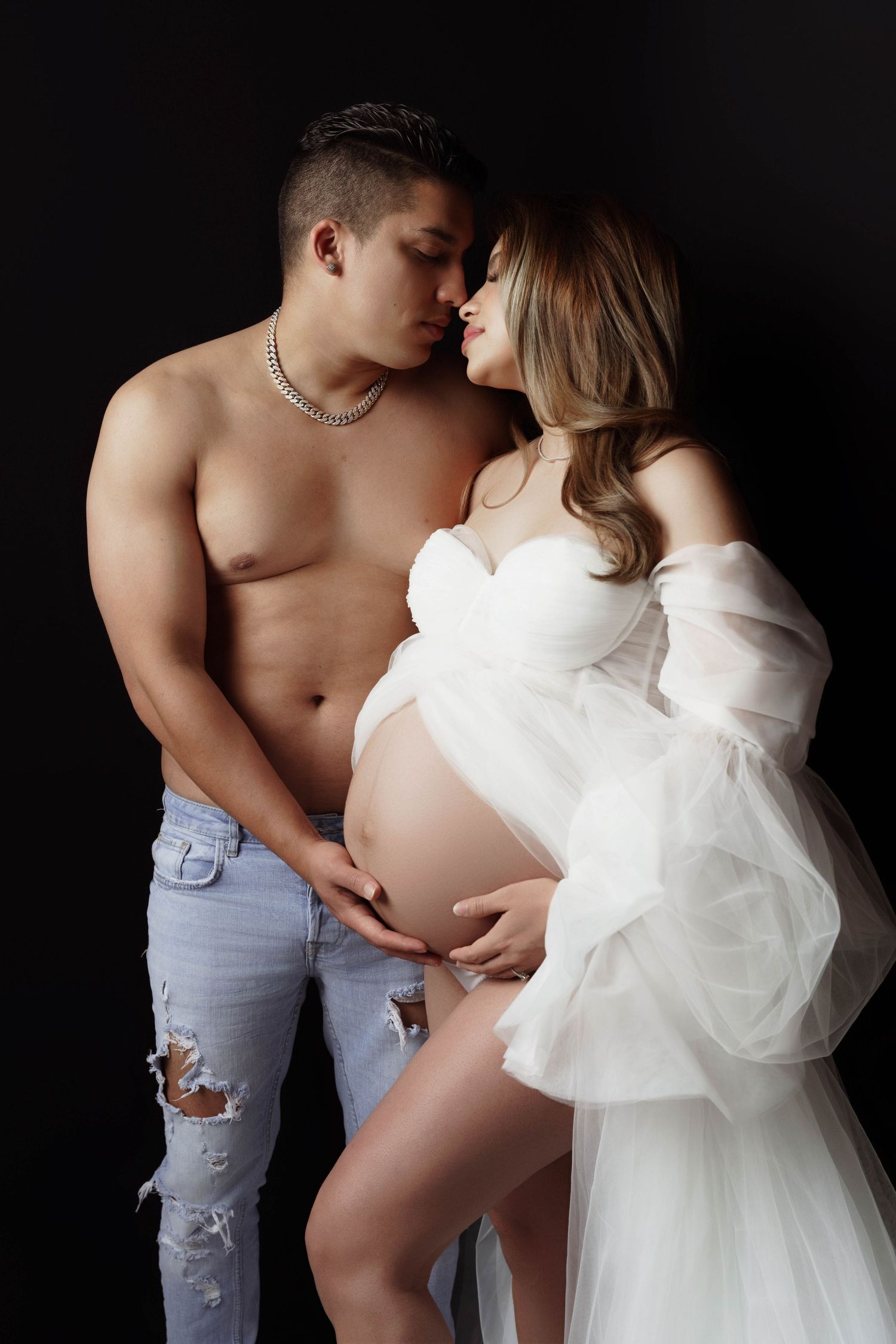 Romantic maternity portraits with couple in white and denim against dark background.