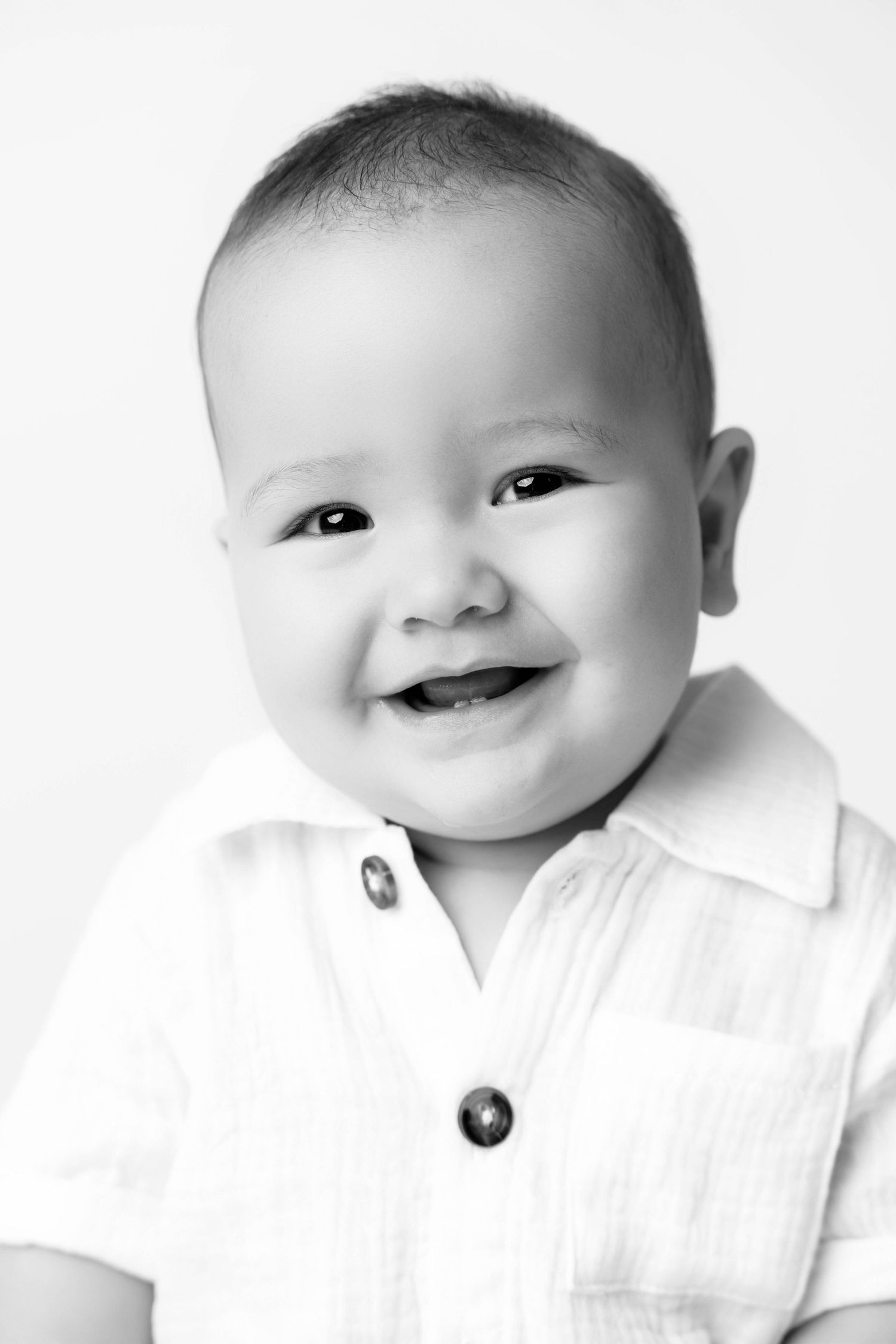 Black and white portrait of a smiling baby boy wearing a white button-up shirt.