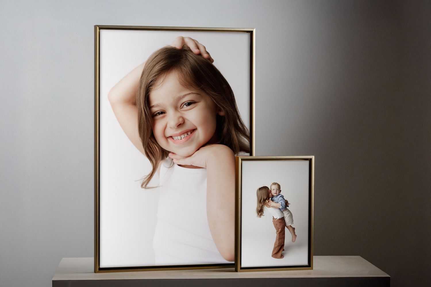 Two gold-framed portrait photos of children displayed on a shelf against a gray wall.