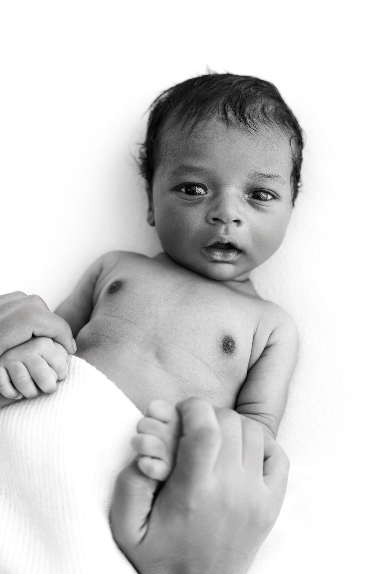 Black and white close-up portrait of a newborn baby lying on white bedding with hands held near their face.