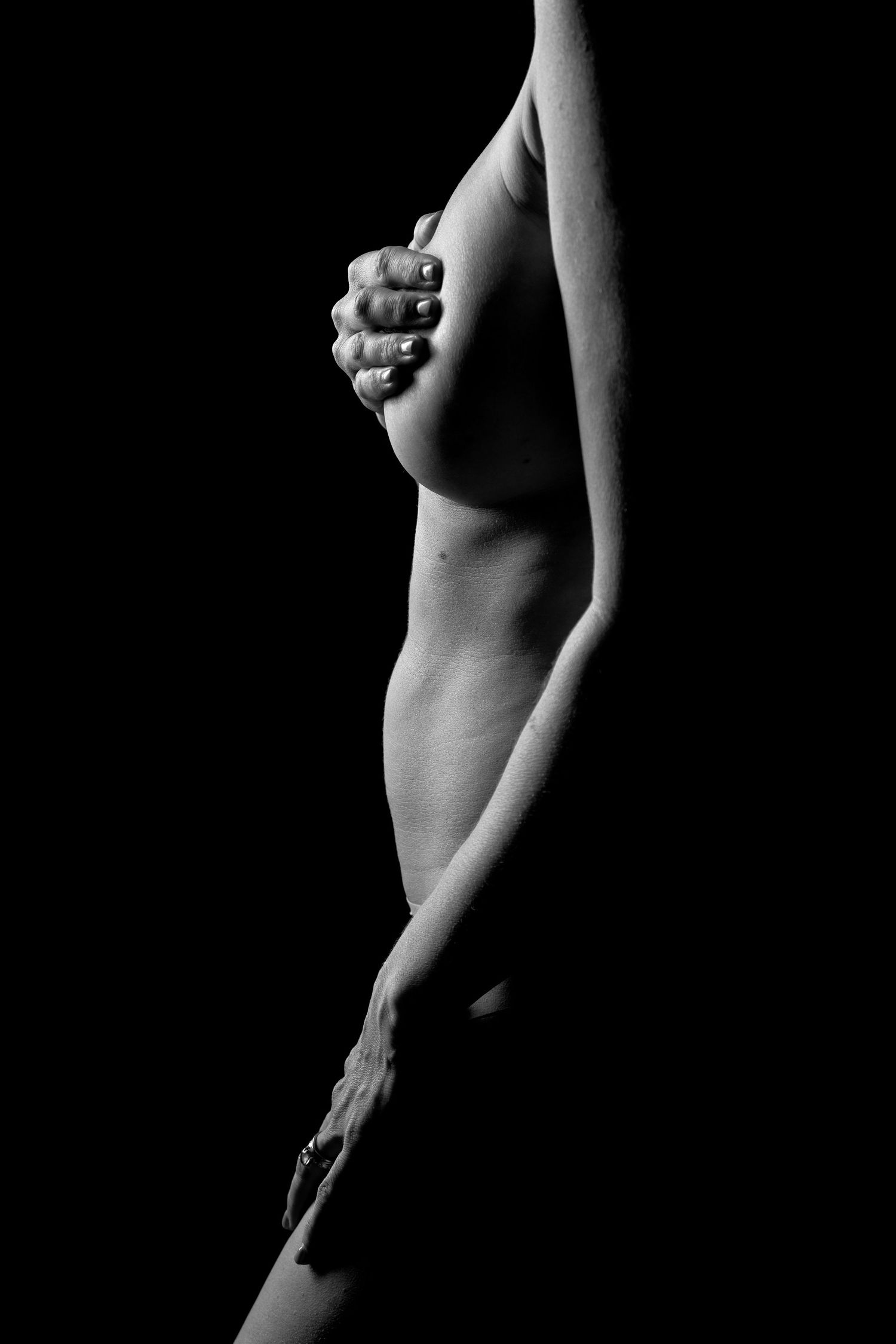 Black and white artistic nude photograph highlighting the female form with dramatic low-key lighting against dark background.