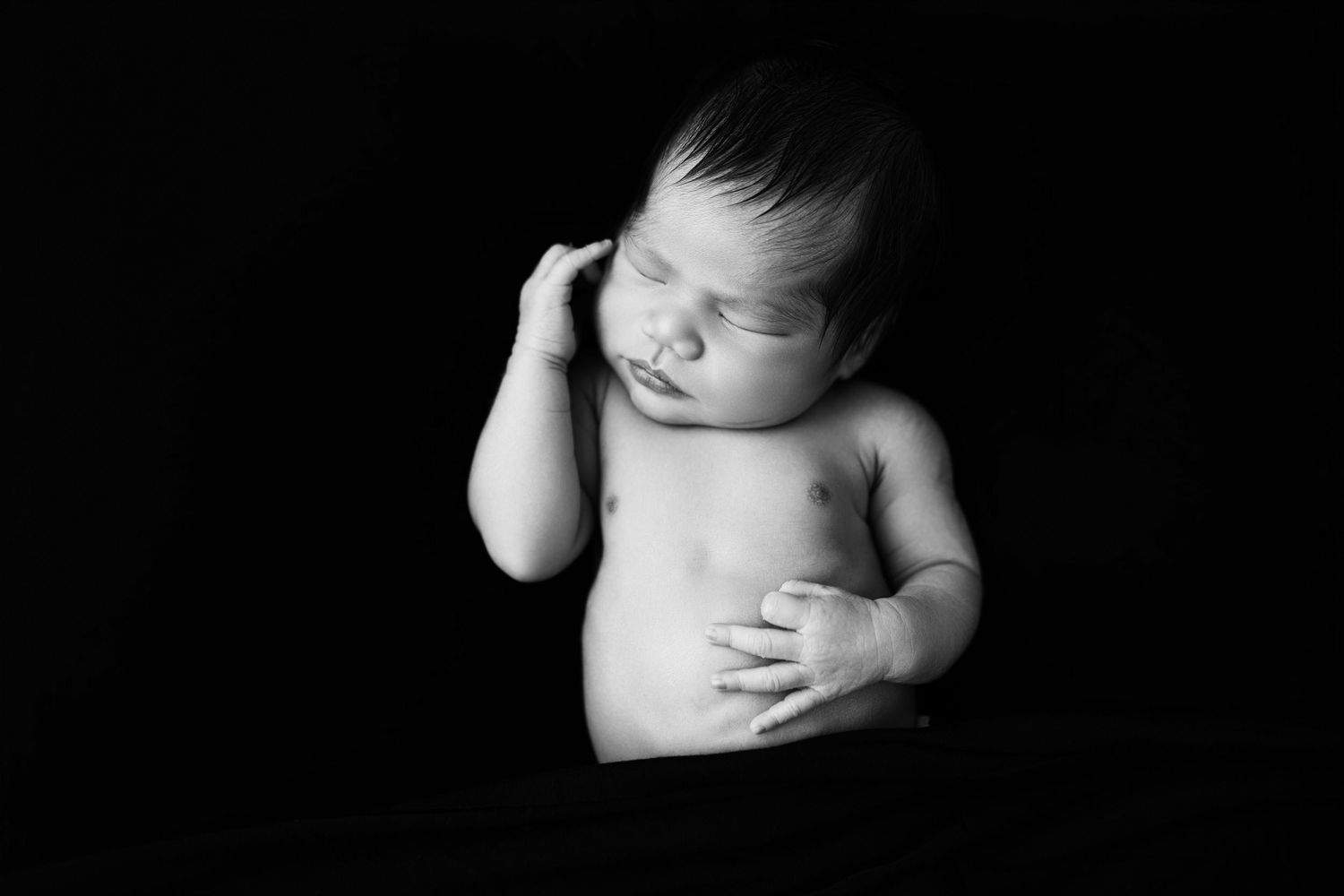 Black and white newborn portrait of a sleeping baby with hand raised to cheek against dark background.