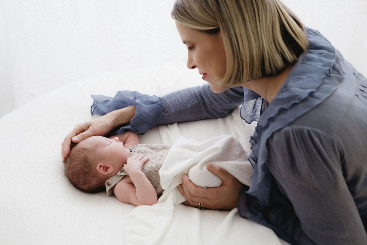A parent in a blue shirt gently interacts with a baby lying on white bedding.