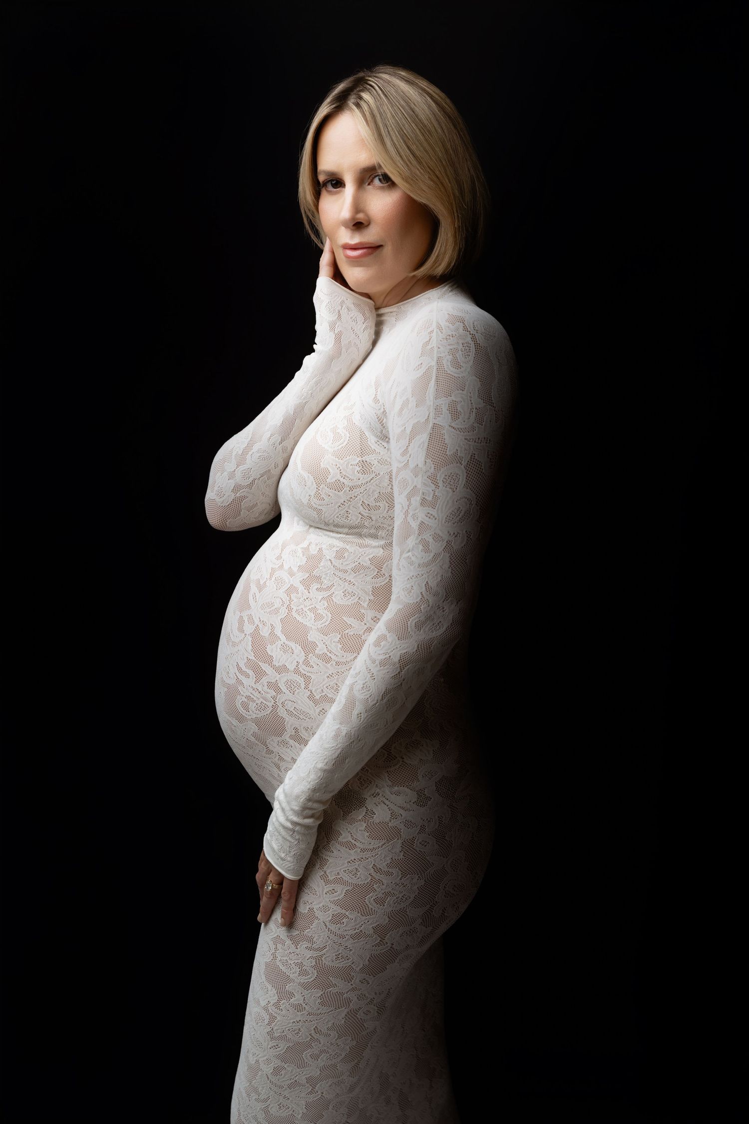 A figure in a white lace maternity dress poses against a black background in an elegant studio portrait series.