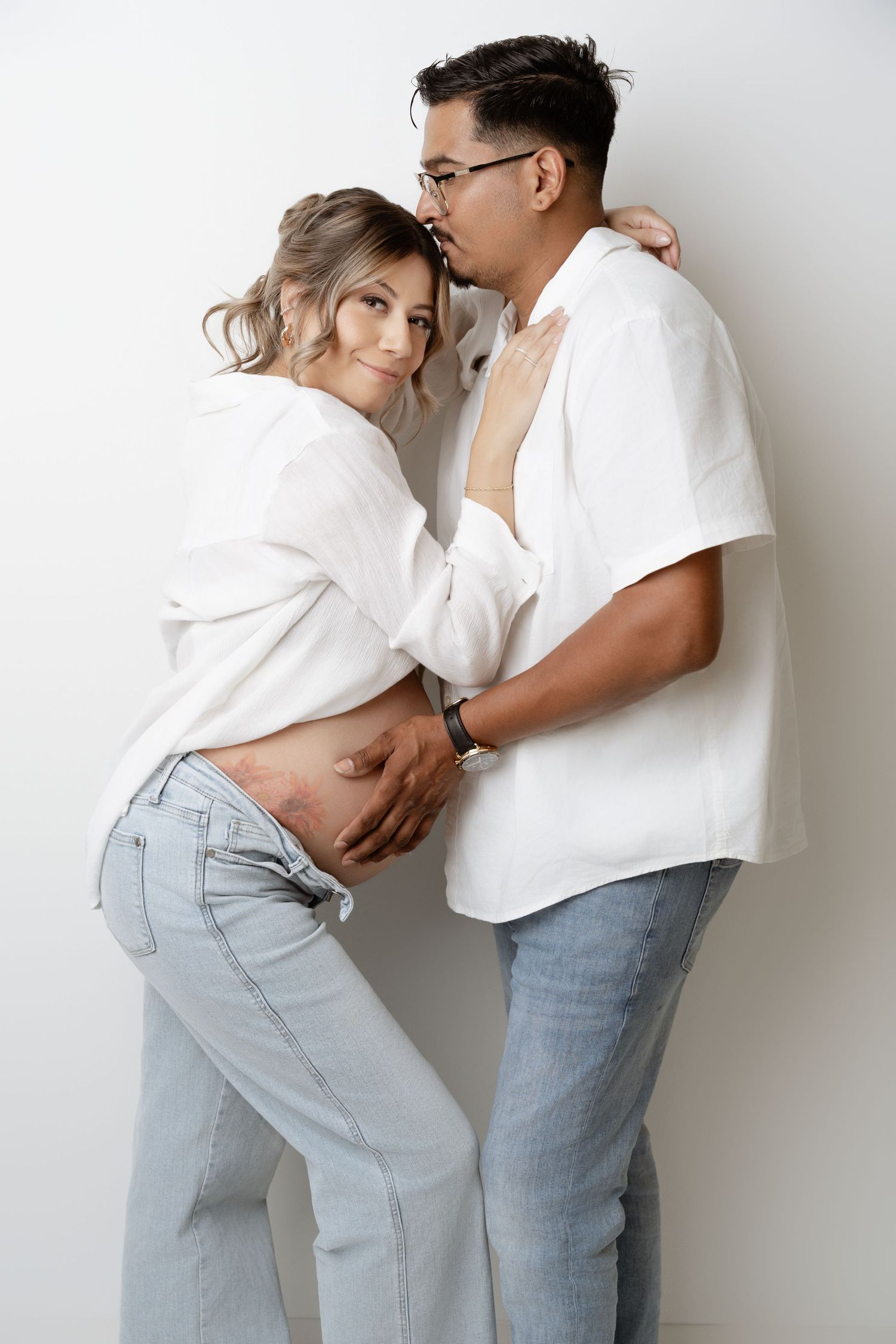 Couple in matching white tops and light jeans embracing against white wall in minimalist maternity photo series.