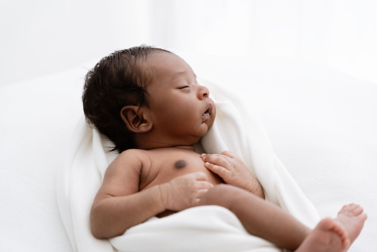 Newborn baby sleeping peacefully on soft white bedding with their arms outstretched.