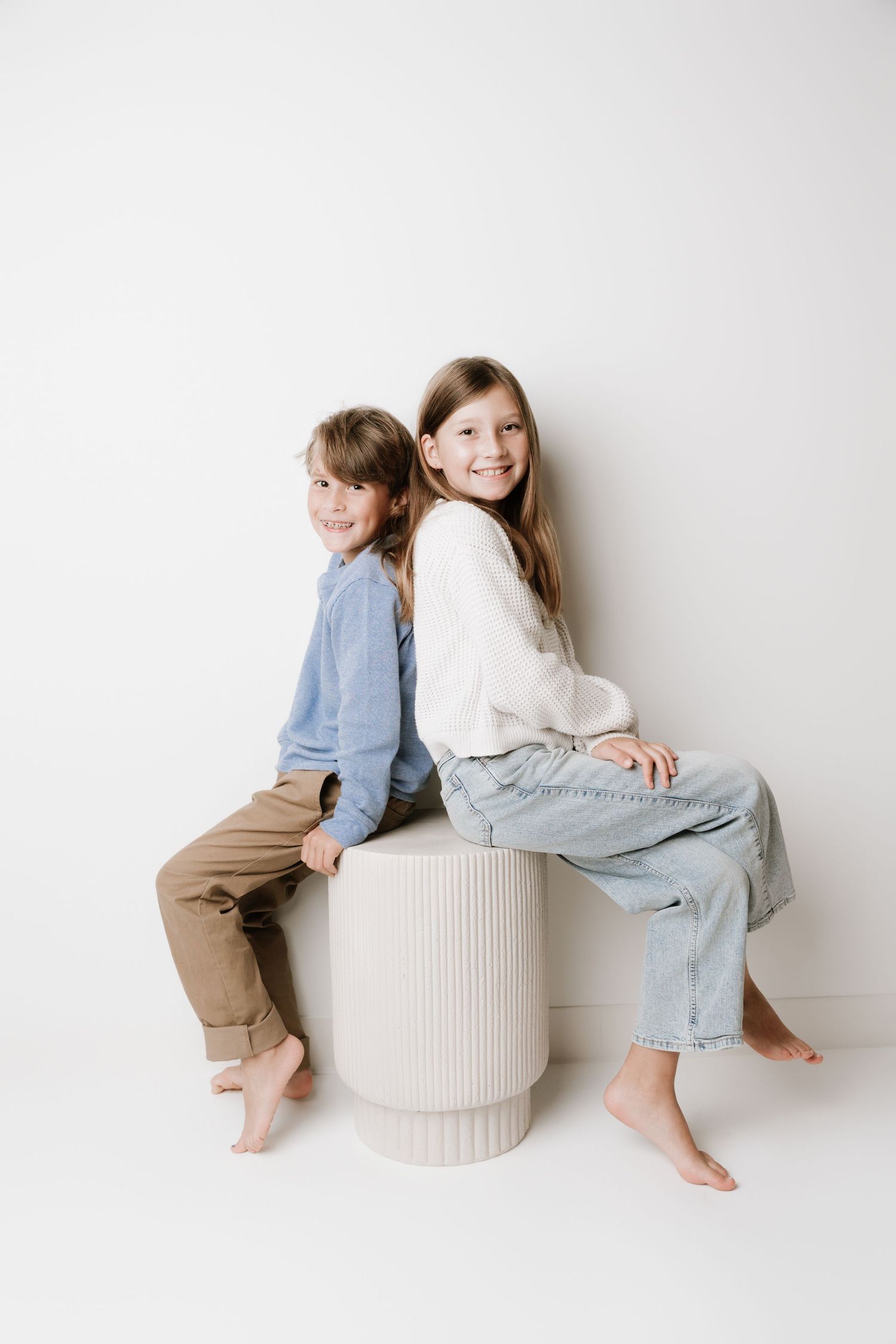Two smiling children sitting back-to-back on a white pedestal stool against a white background.