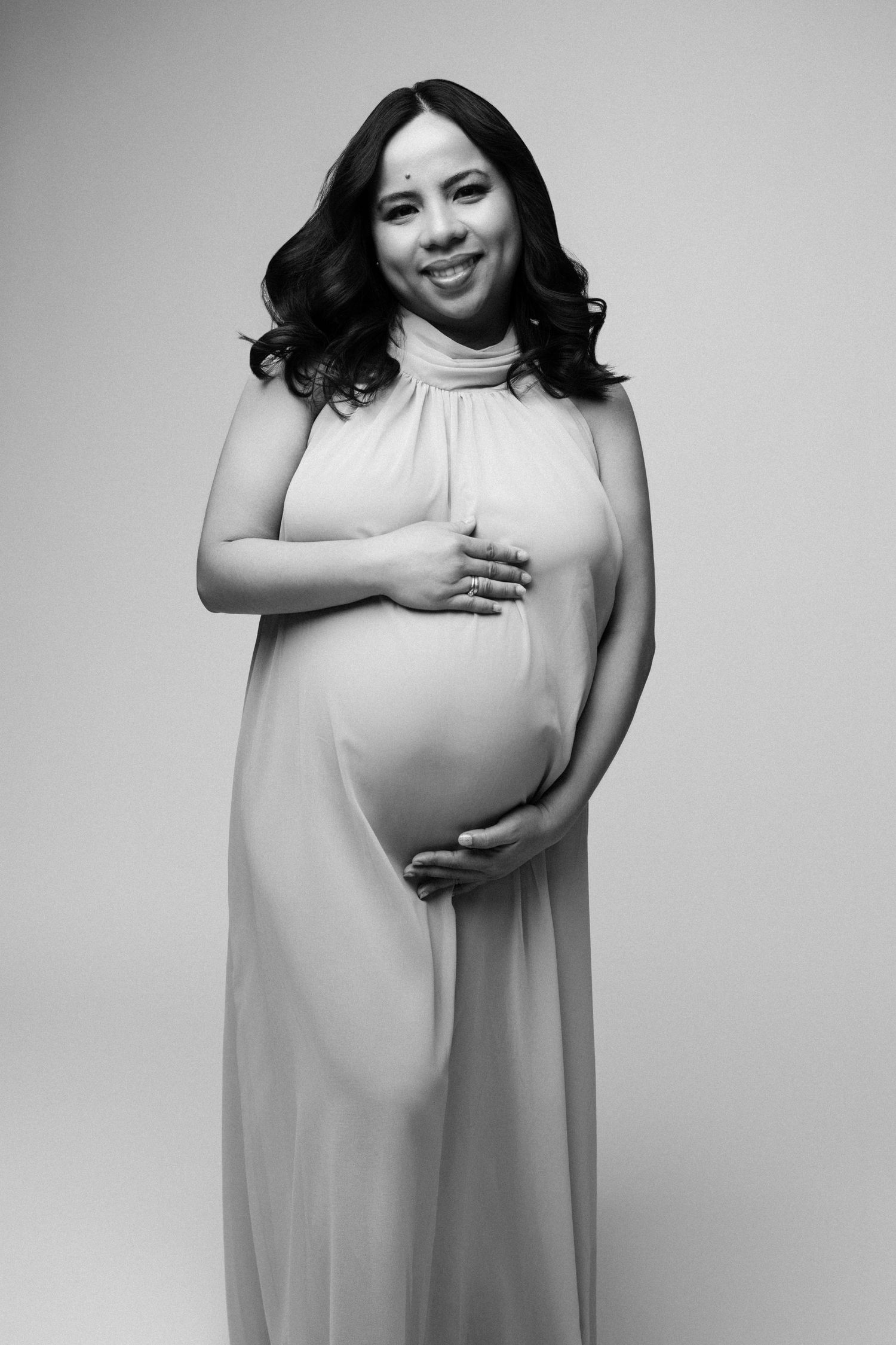 Black and white maternity portrait of an expectant mother in a flowing white dress against a light background.