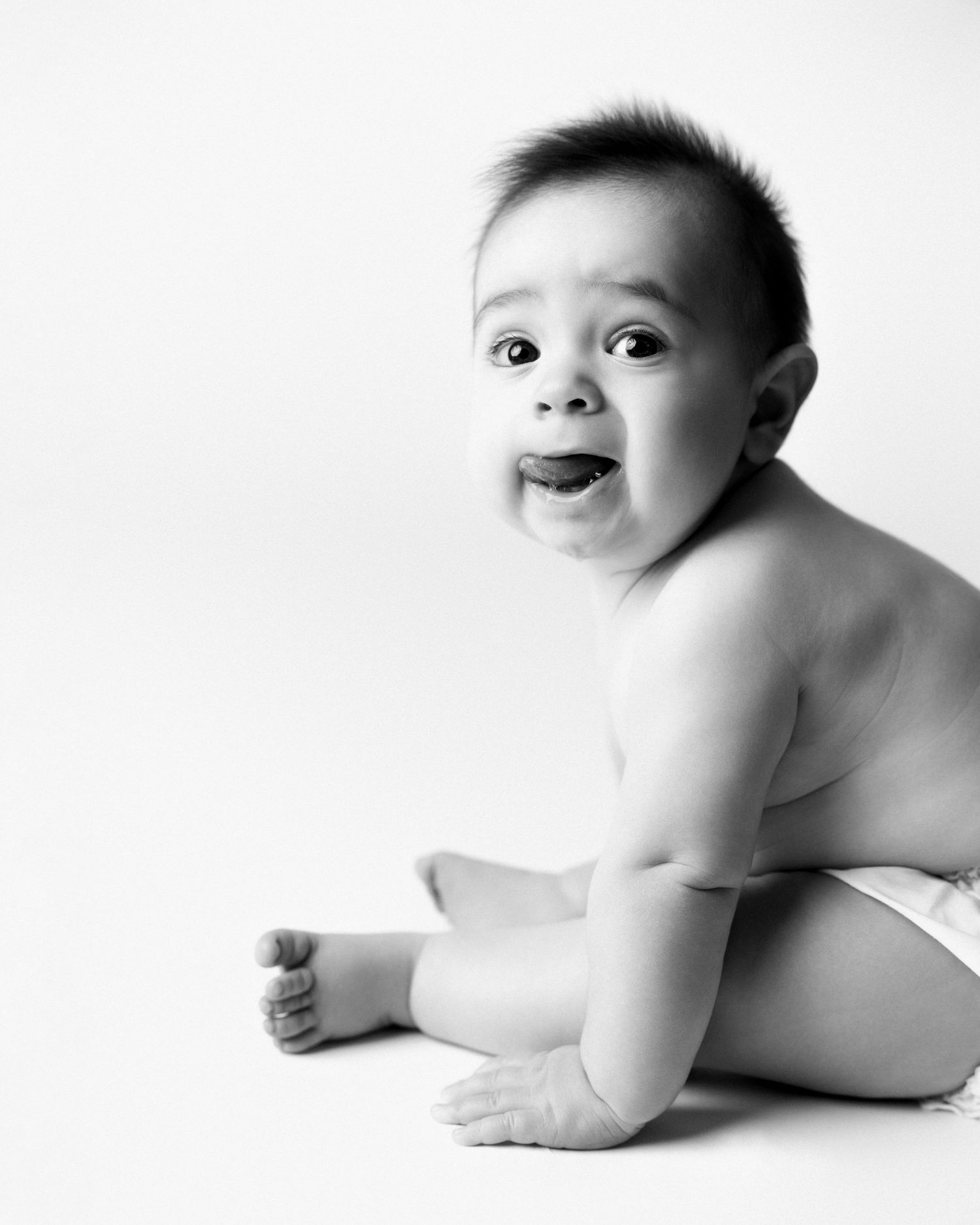 Black and white photo of an adorable baby sitting up, wearing a diaper, with a cute surprised expression.
