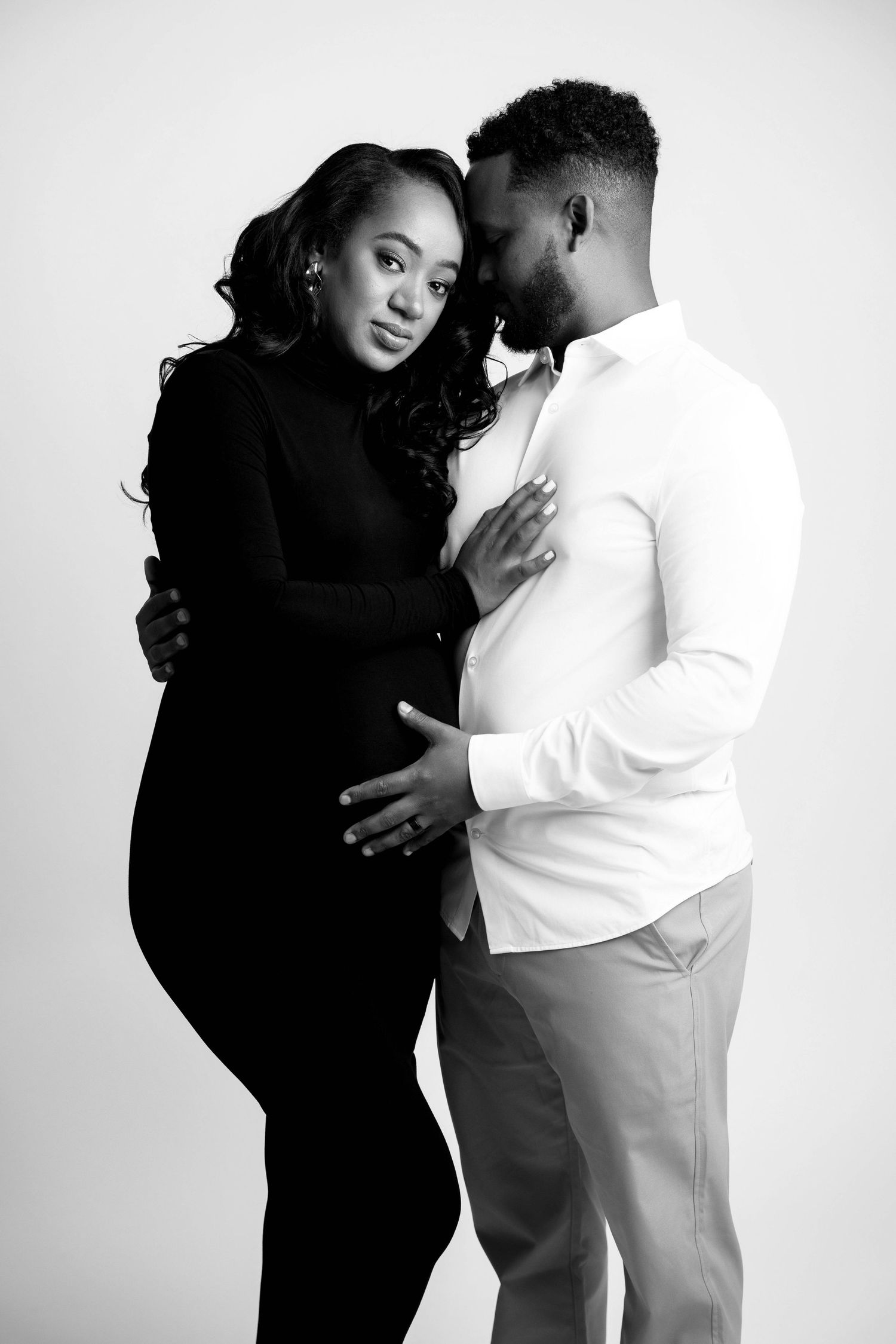 Romantic maternity portraits of couple in black and white with woman in dark dress against light background.