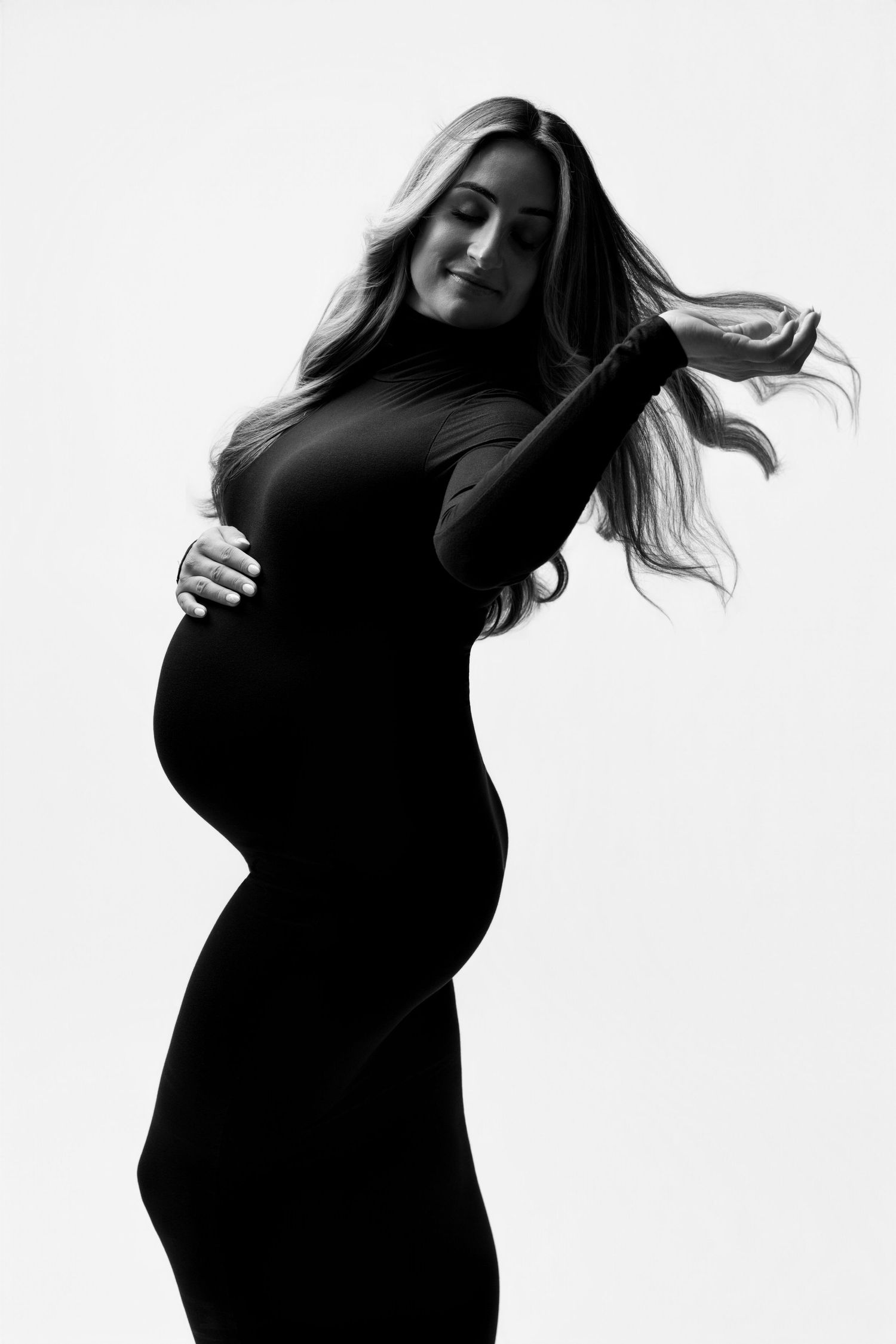 Black and white maternity photo of a pregnant woman in a black dress cradling her baby bump with flowing hair.