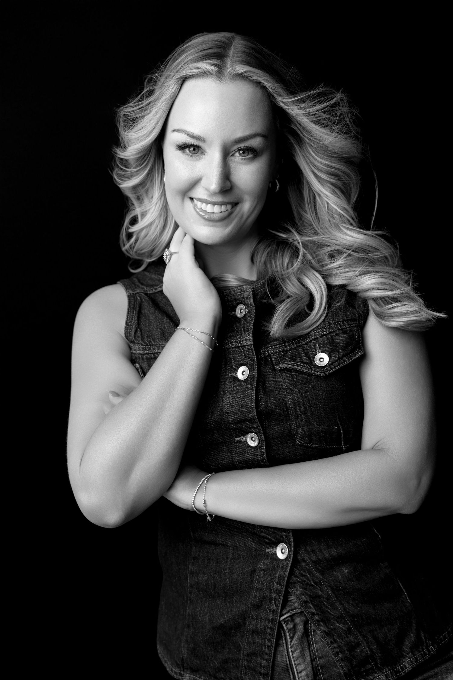 Black and white portrait of a smiling blonde woman in a denim vest posing against a dark background.