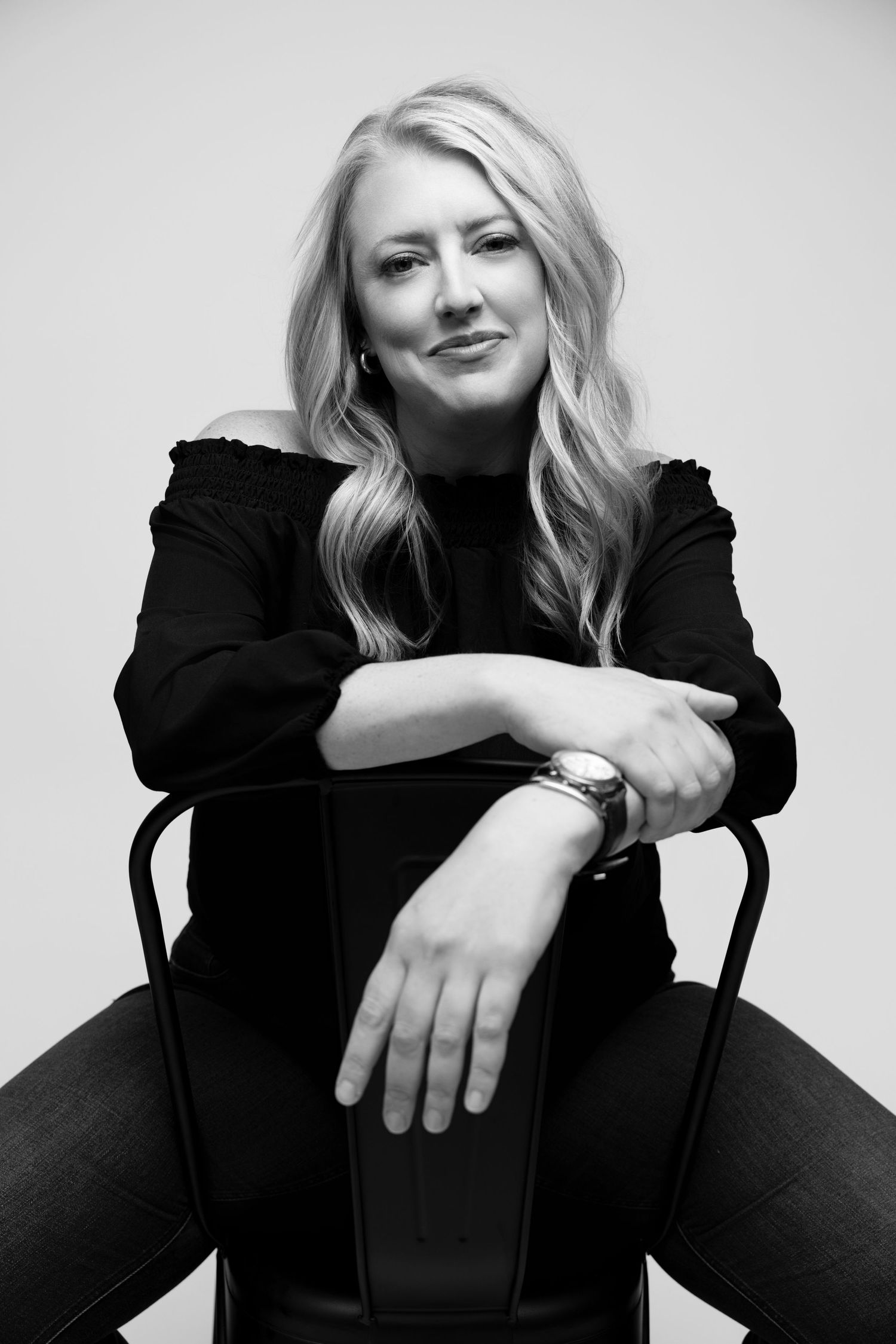 Black and white portrait of a blonde woman in a black off-shoulder top, seated on a chair, smiling confidently.