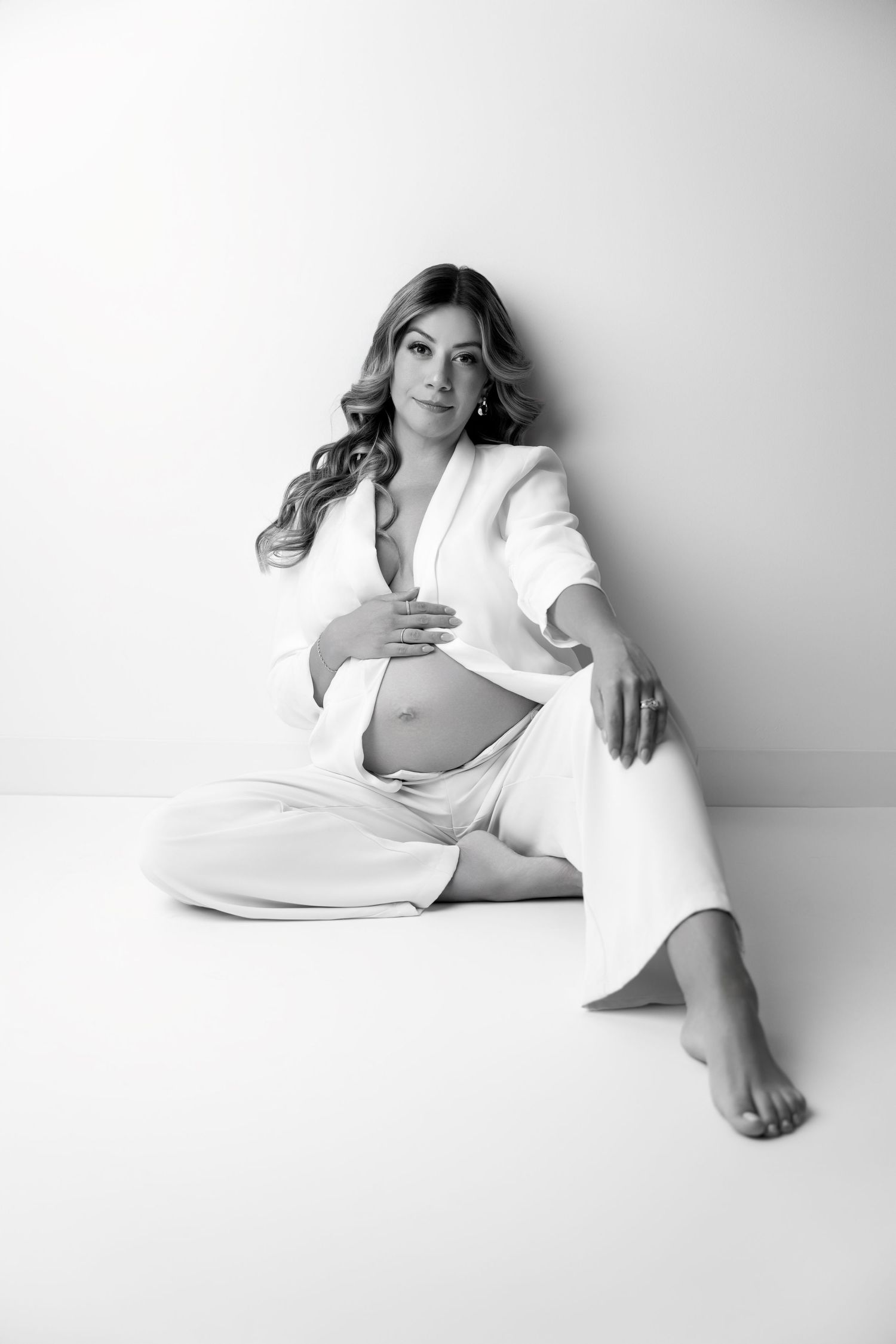 Black and white maternity portrait of someone in a white outfit sitting on floor against white backdrop in elegant pose.