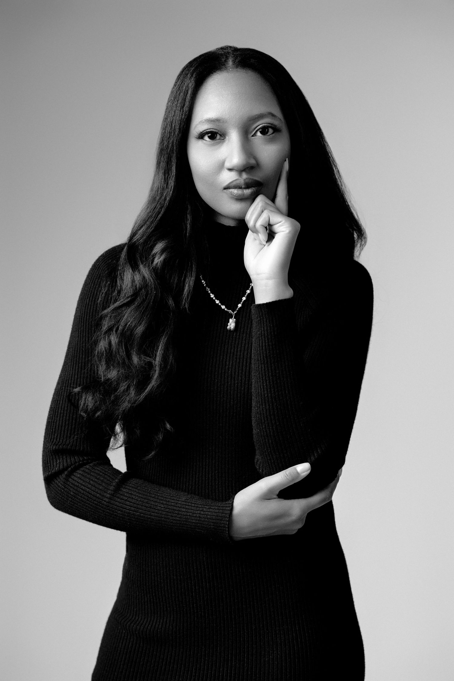Black and white portrait of a young woman in a turtleneck sweater, posing thoughtfully with hand on chin.