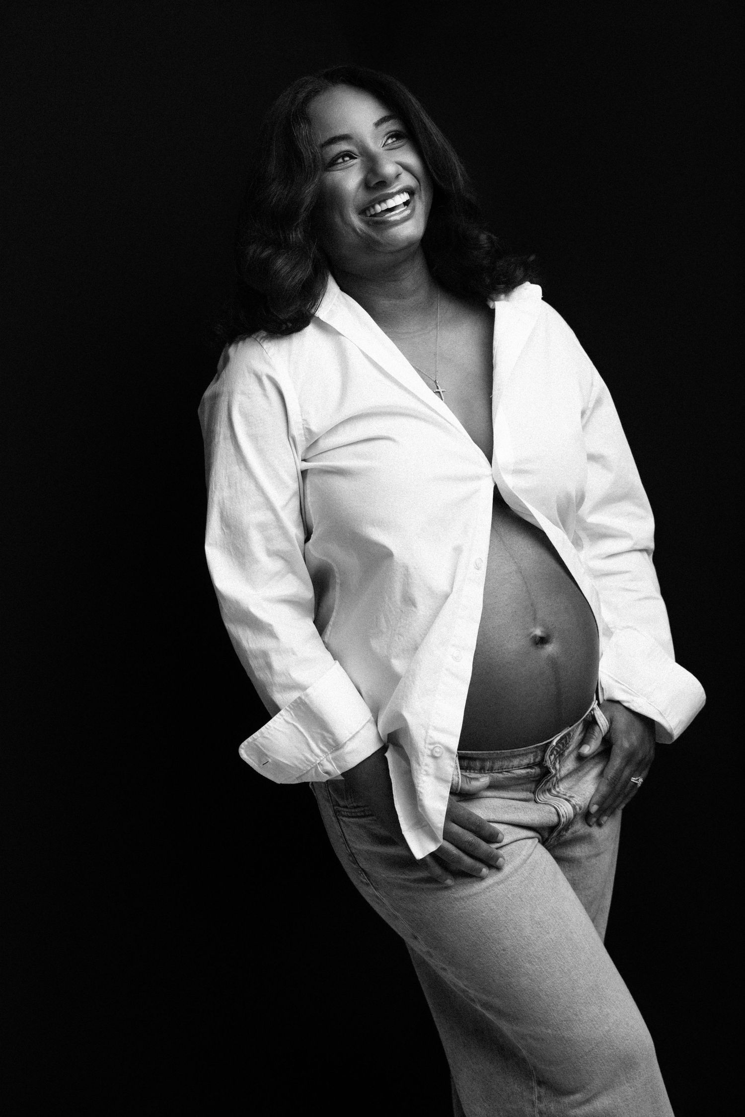 Black and white portrait of a smiling pregnant woman in an open white shirt and jeans, showcasing her baby bump.